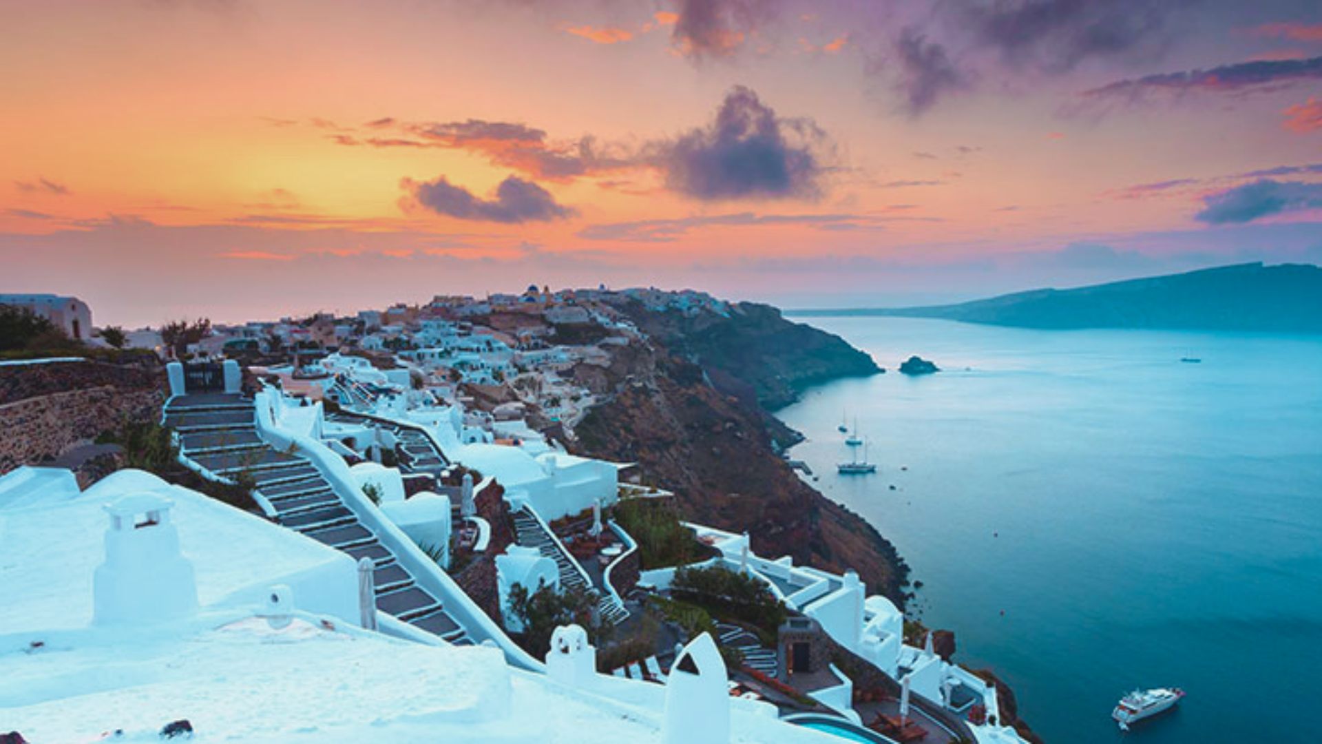 Sunset view of Santorini, Greece, with white cliffside buildings overlooking a calm, blue sea. The sky is painted with warm hues, conveying a serene ambiance.