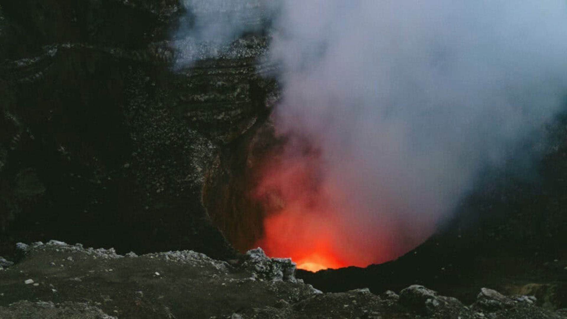 A fiery red glow emanates from the depths of an active volcano crater, shrouded in thick white smoke under a dark, rocky landscape, conveying intense natural power.