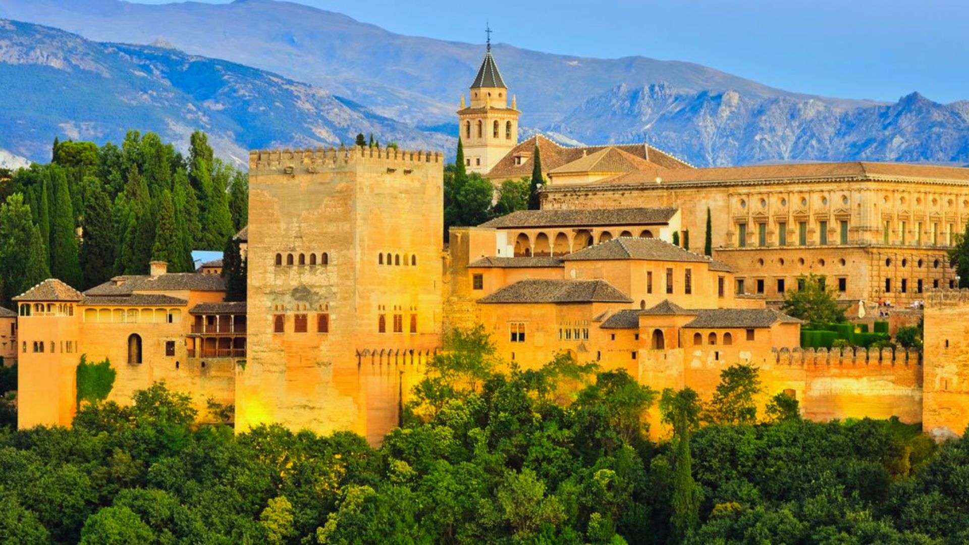 A majestic view of the Alhambra in Granada, Spain, with its historic, sunlit stone architecture set against a backdrop of lush greenery and distant mountains.