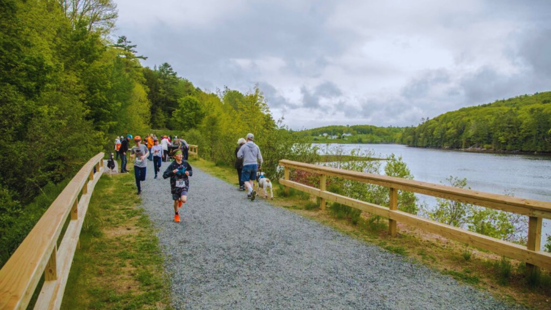 A group of people walk and jog on a gravel path beside a serene lake, surrounded by lush green trees. The scene is calm and lively under a cloudy sky.