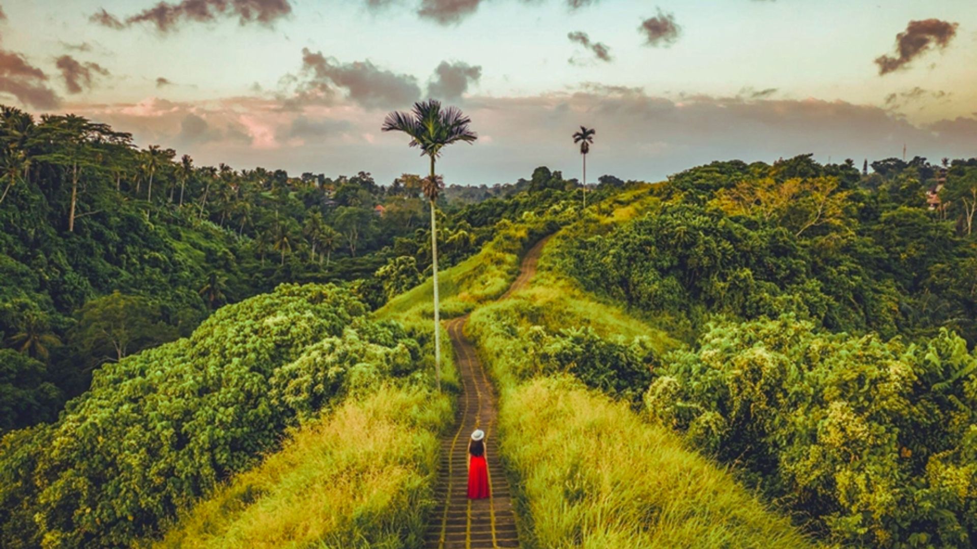 A lone person in a red dress and hat walks along a narrow path amid lush green hills under a pastel-colored sky, conveying serenity and adventure.