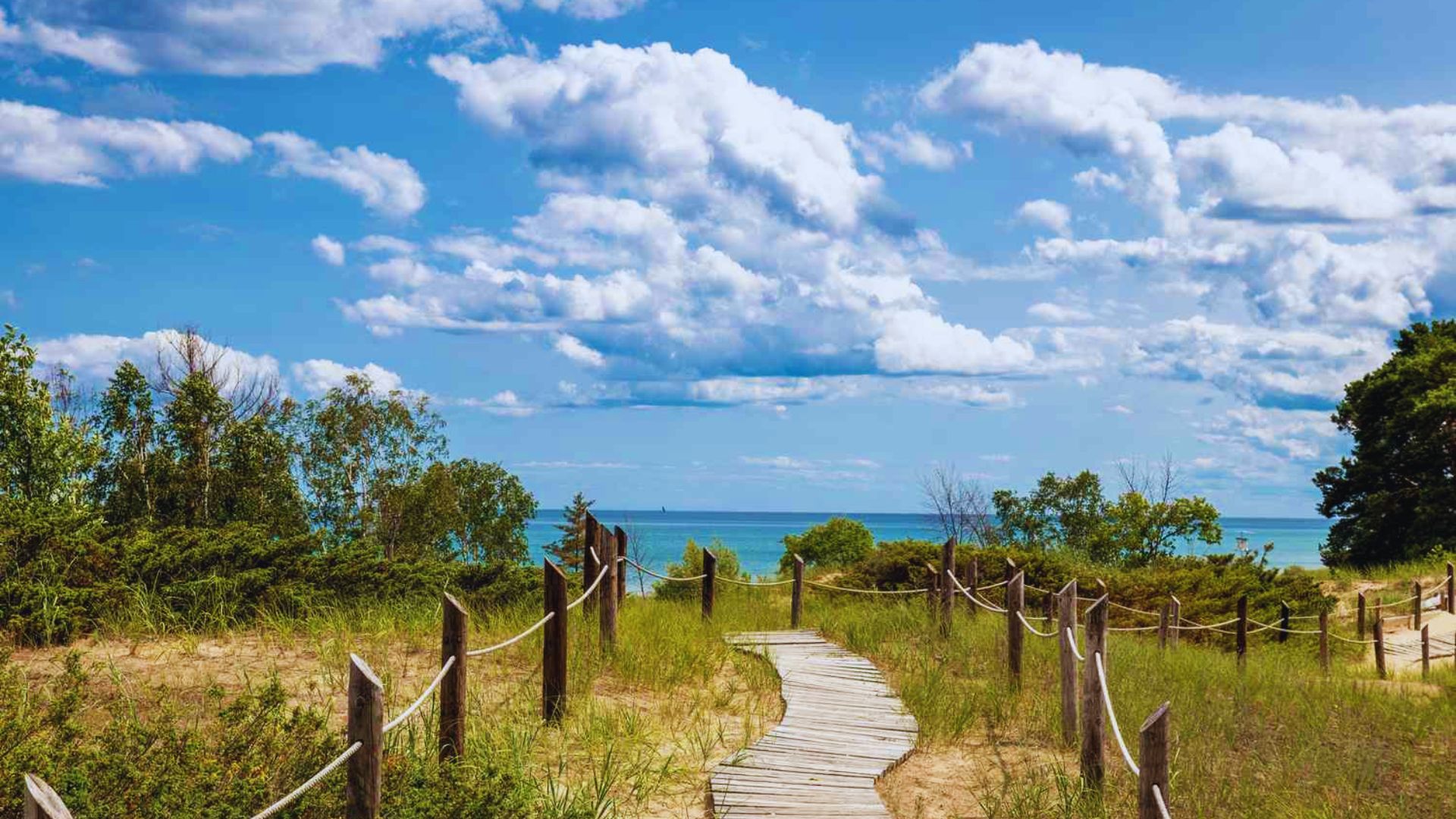 Pathway through grassy dunes leads to a tranquil beach with turquoise water. The sky is bright blue with fluffy clouds, conveying peacefulness.
