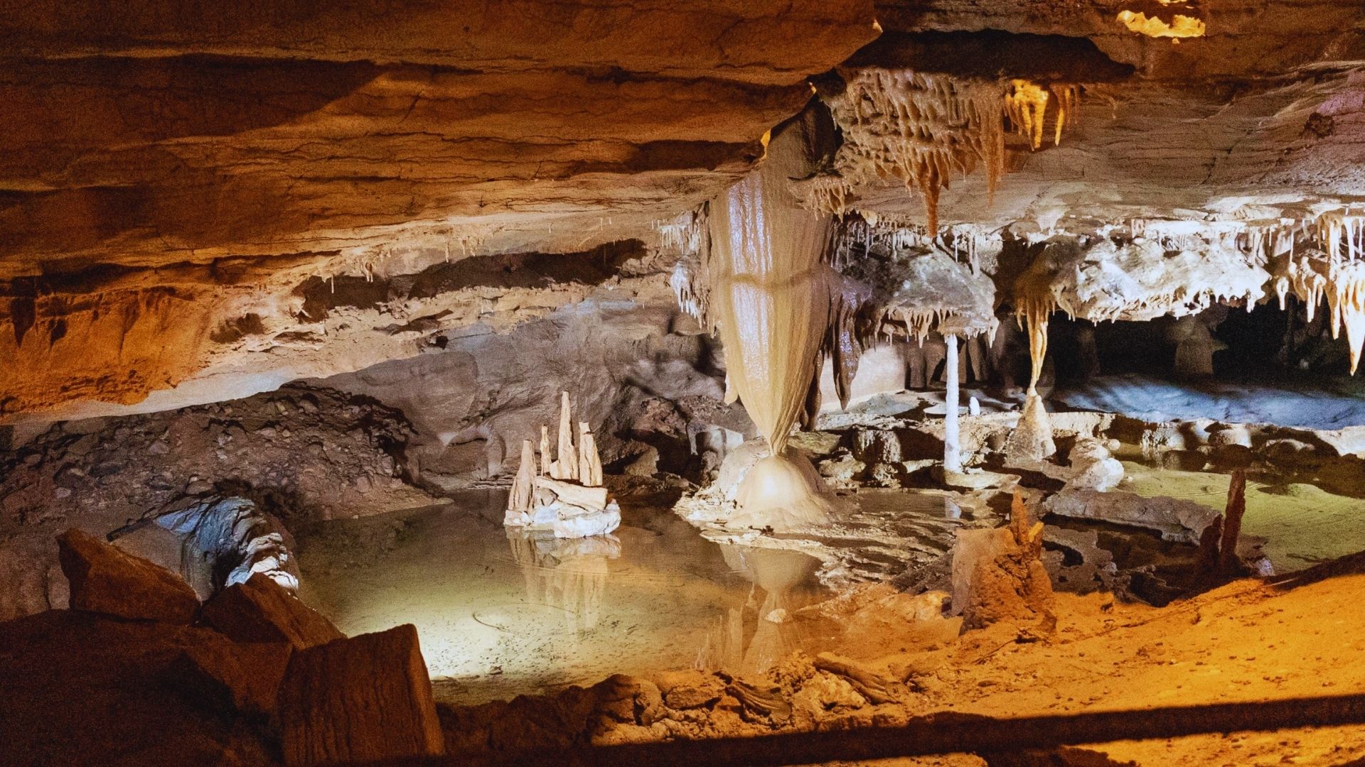 A dimly lit cave with majestic stalactites and stalagmites reflected in a calm pool of water. The ambiance is serene and mysterious.