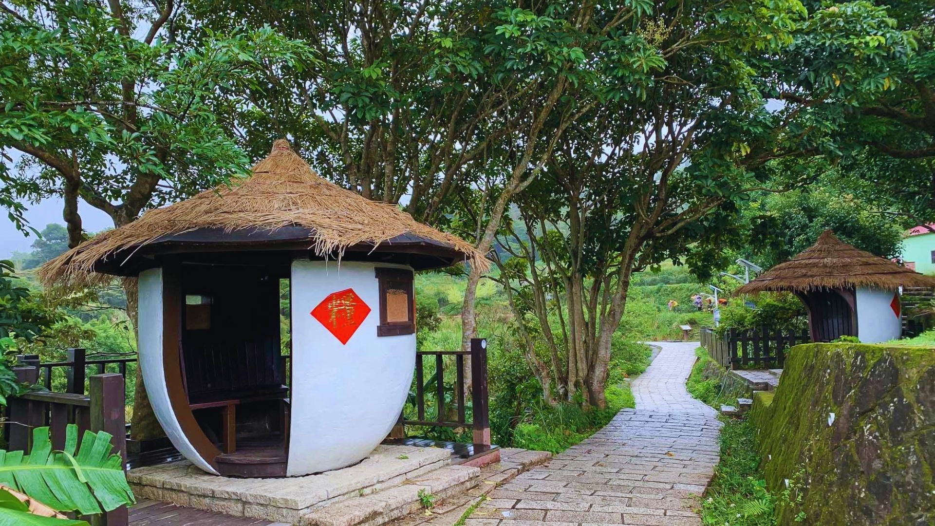 Small, white, round hut with thatched roof beside a stone path winding through lush greenery and trees. Peaceful, rustic, and inviting scene.