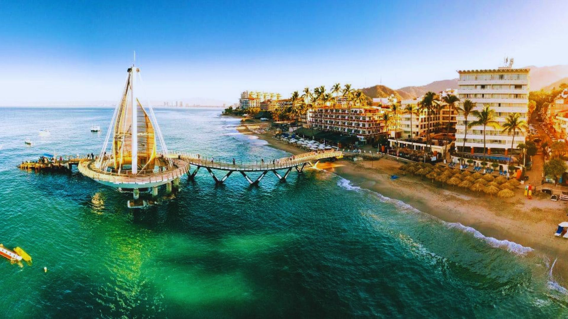 A scenic view of a beachside city with a modern pier extending into turquoise waters. Lined with palm trees, the shoreline features umbrellas and beachfront hotels under a clear blue sky.