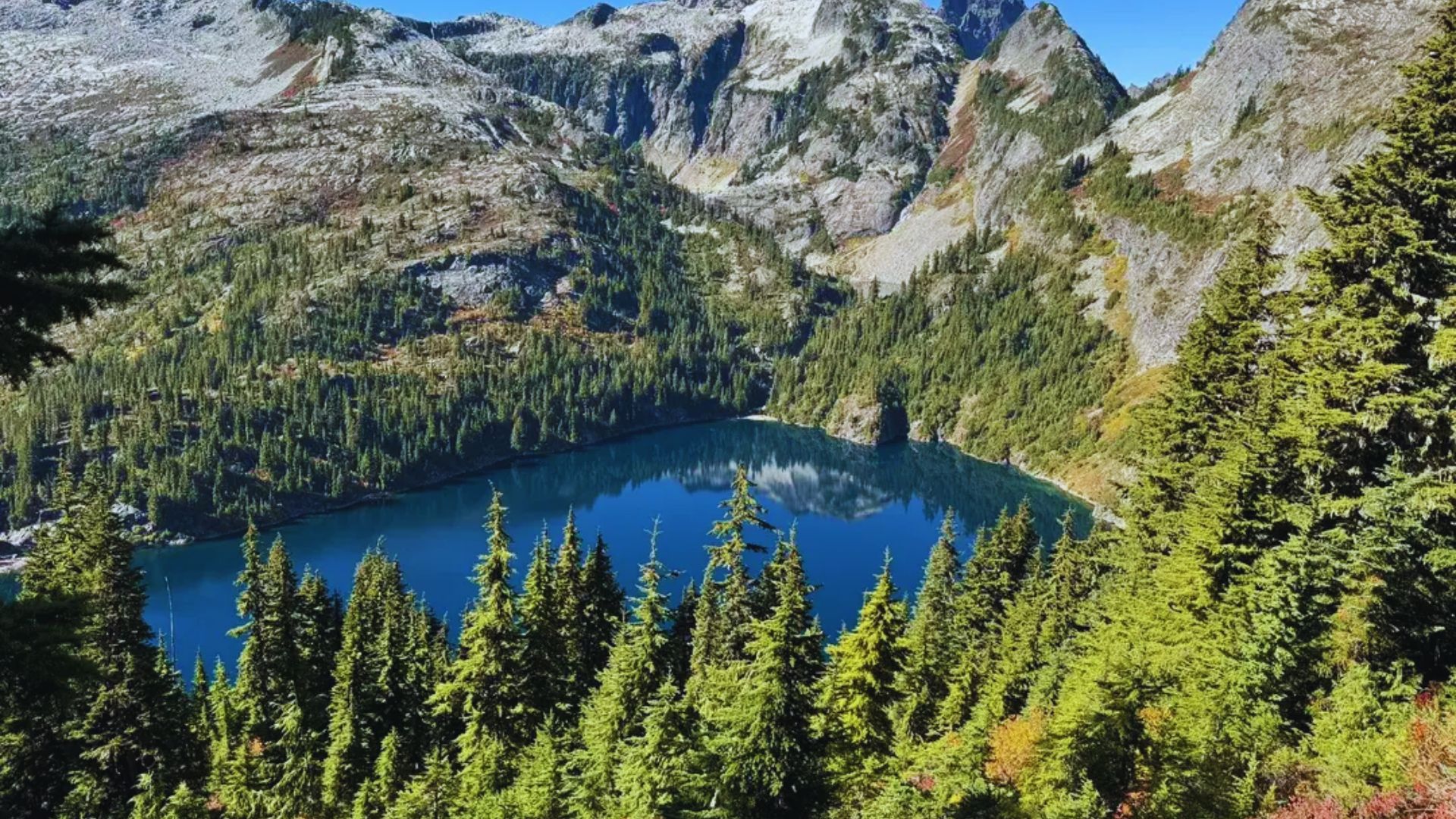 A serene mountain lake reflects a clear blue sky, surrounded by lush evergreen trees and rugged mountains. The view is tranquil and picturesque.