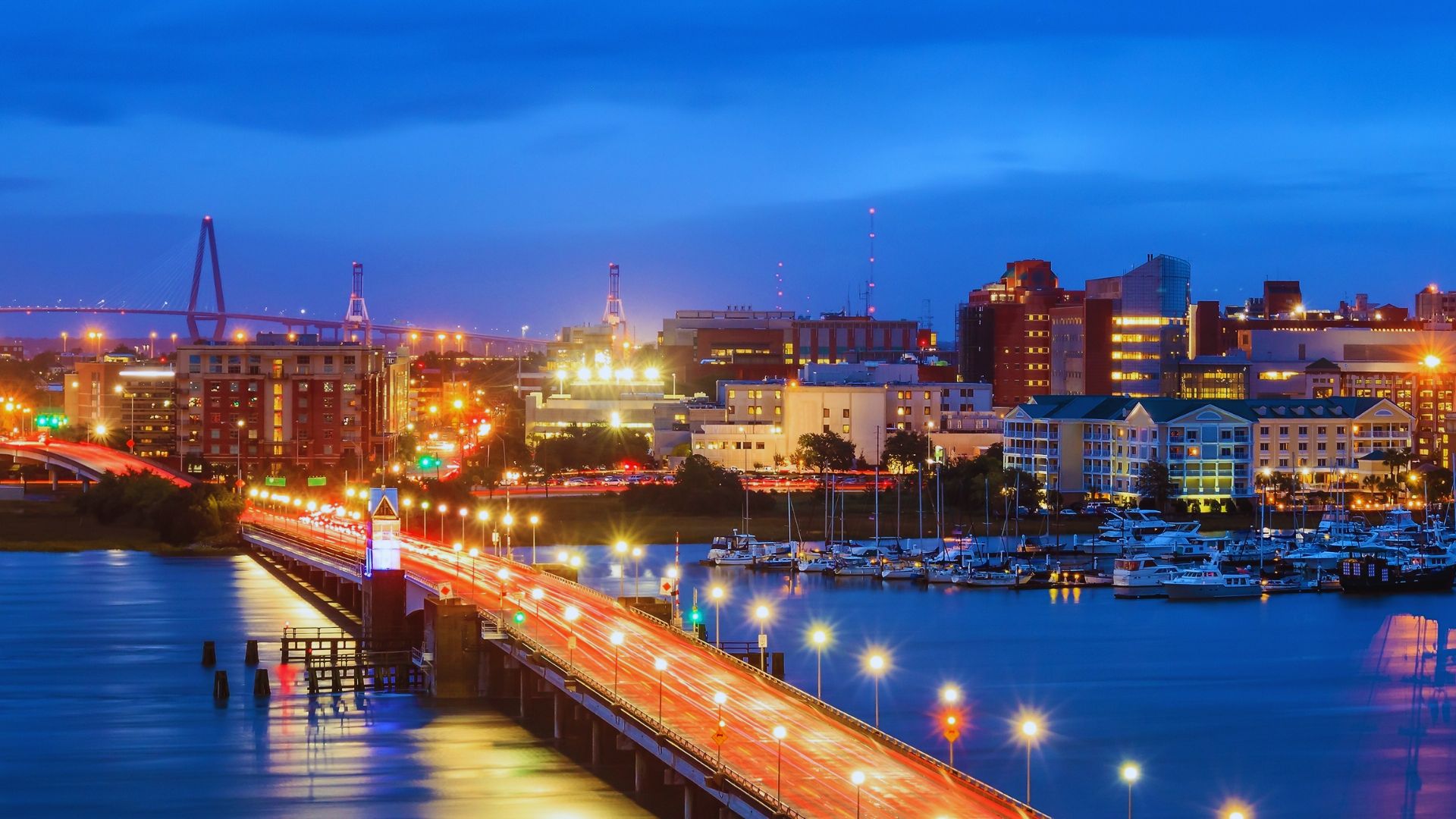 Nighttime cityscape featuring a lit-up bridge leading to a vibrant urban area with colorful city lights, docked boats, and a calm blue sky.