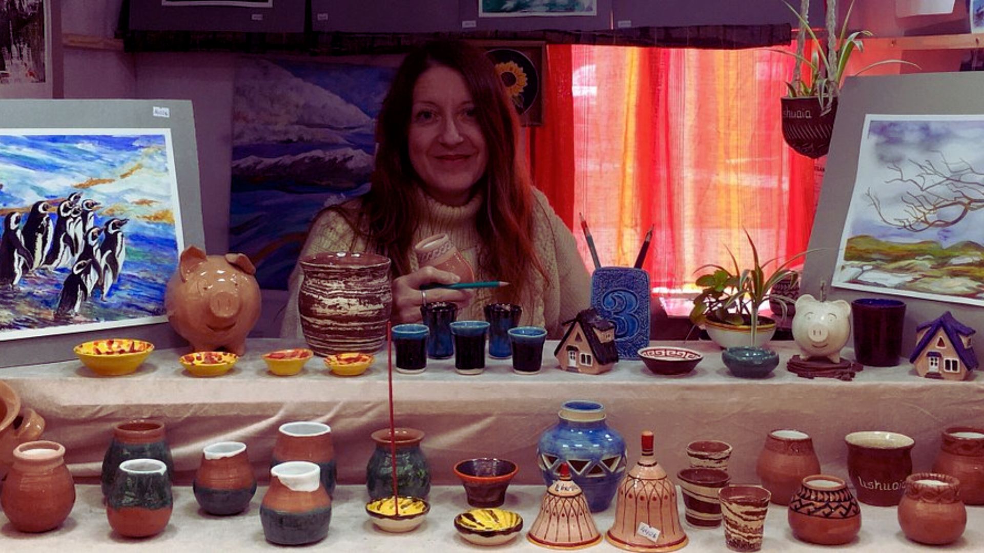 A woman smiles behind a display of colorful handmade pottery and paintings, including jugs, vases, and artwork of penguins. The scene is warm and inviting.