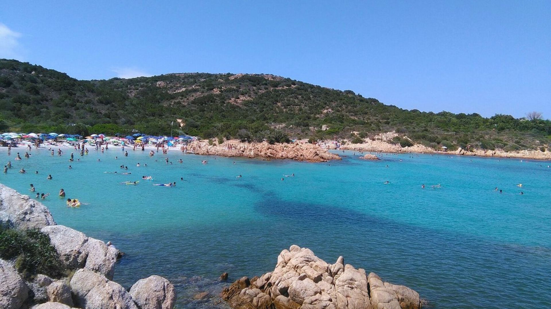 A picturesque beach scene with turquoise waters, rocky shorelines, and lush green hills. People swim and relax under colorful umbrellas, creating a vibrant, inviting atmosphere.