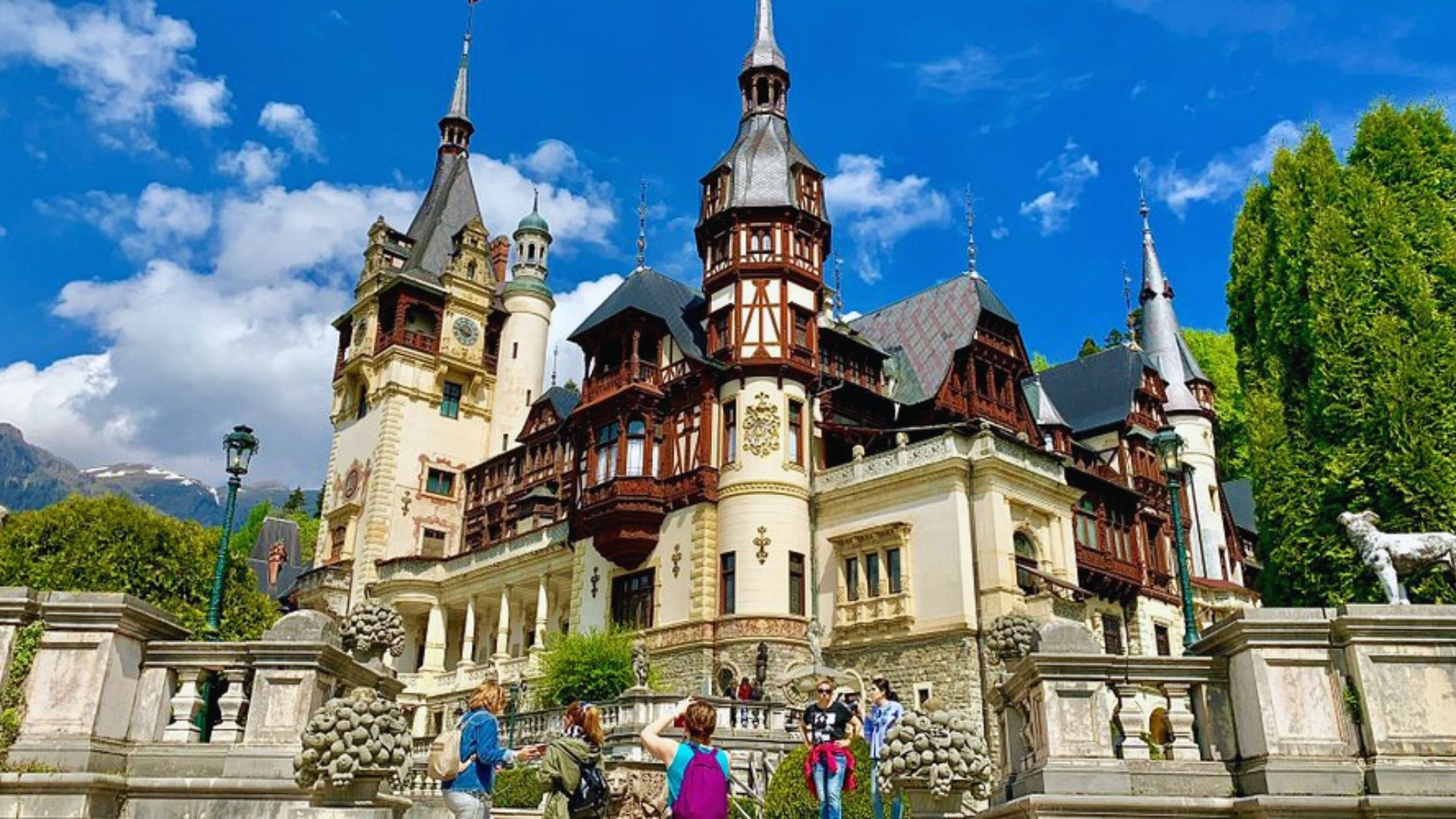 A grand, ornate castle with intricate towers and pointed roofs set against a bright blue sky. Visitors explore the lush garden in the foreground.