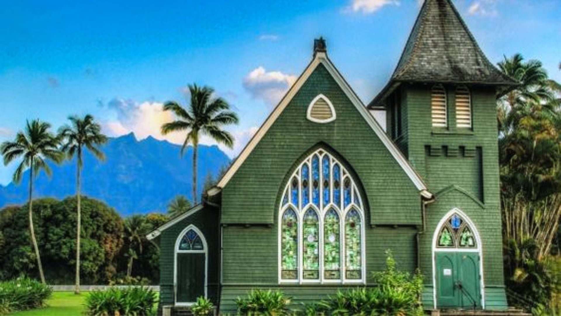 A charming green church with stained-glass windows stands amidst lush greenery and palm trees, with distant mountains under a clear blue sky.