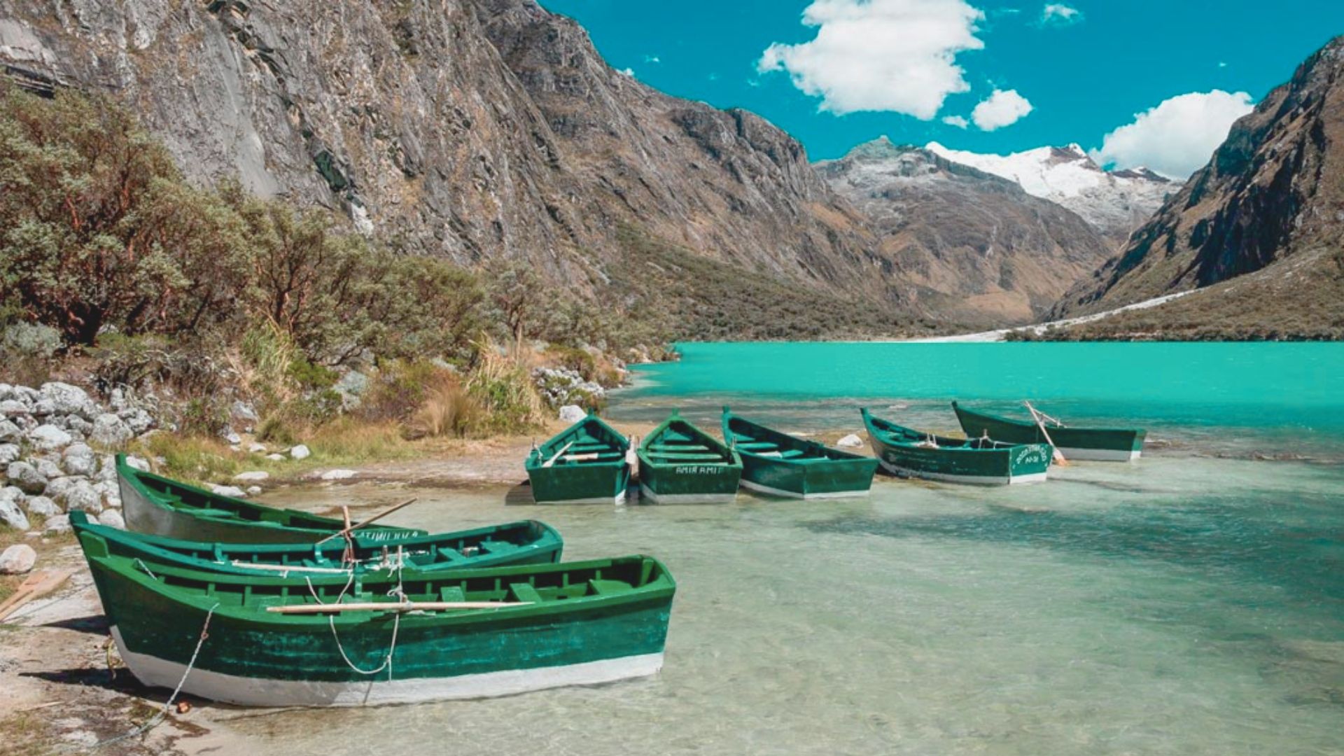 Green rowboats rest on the clear turquoise waters of a serene mountain lake, surrounded by rocky cliffs and distant snow-capped peaks under a bright blue sky.