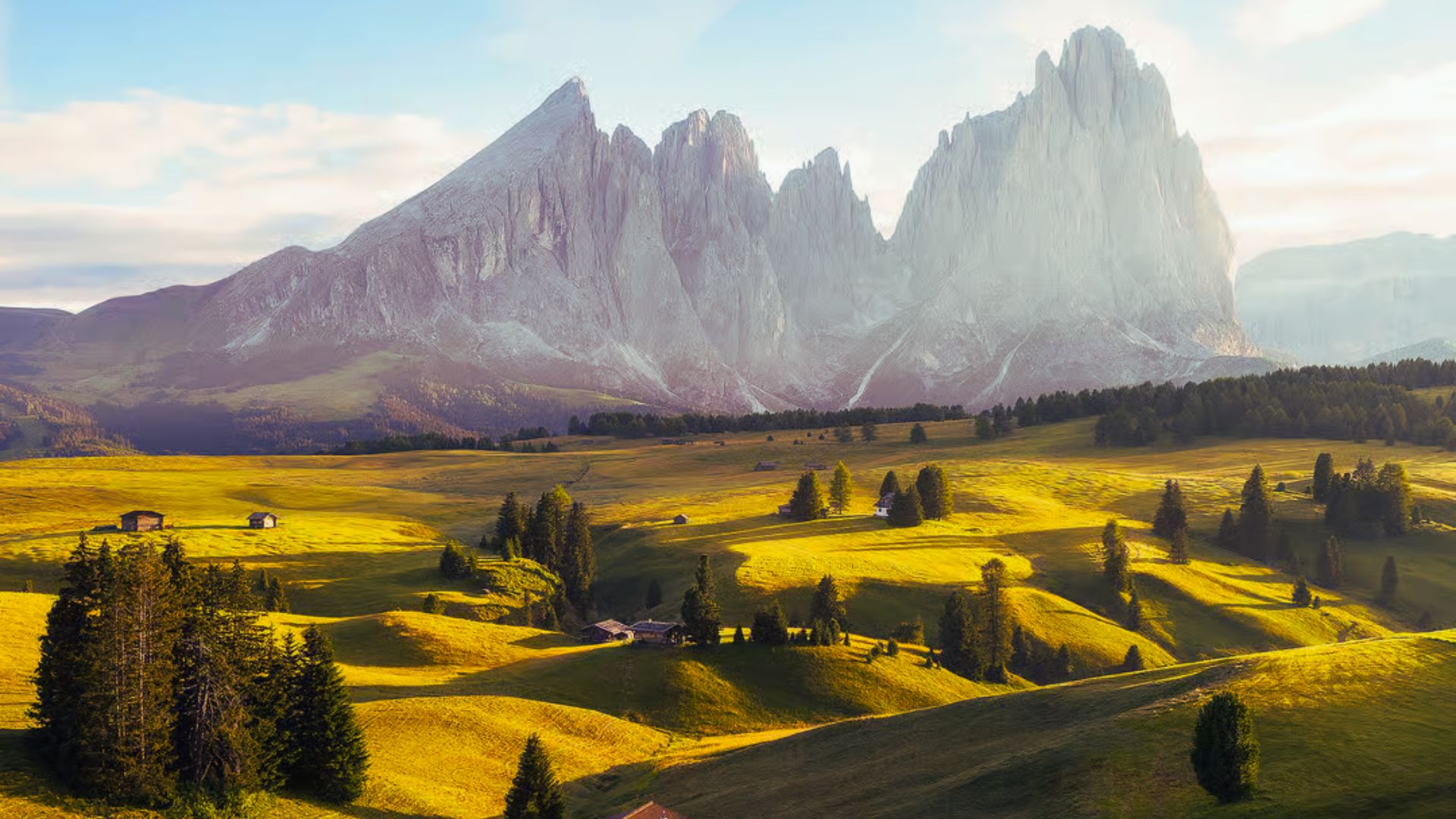 Lush rolling hills bathed in warm sunlight, dotted with trees and small cabins, with majestic, rugged mountains towering in the background under a clear sky.