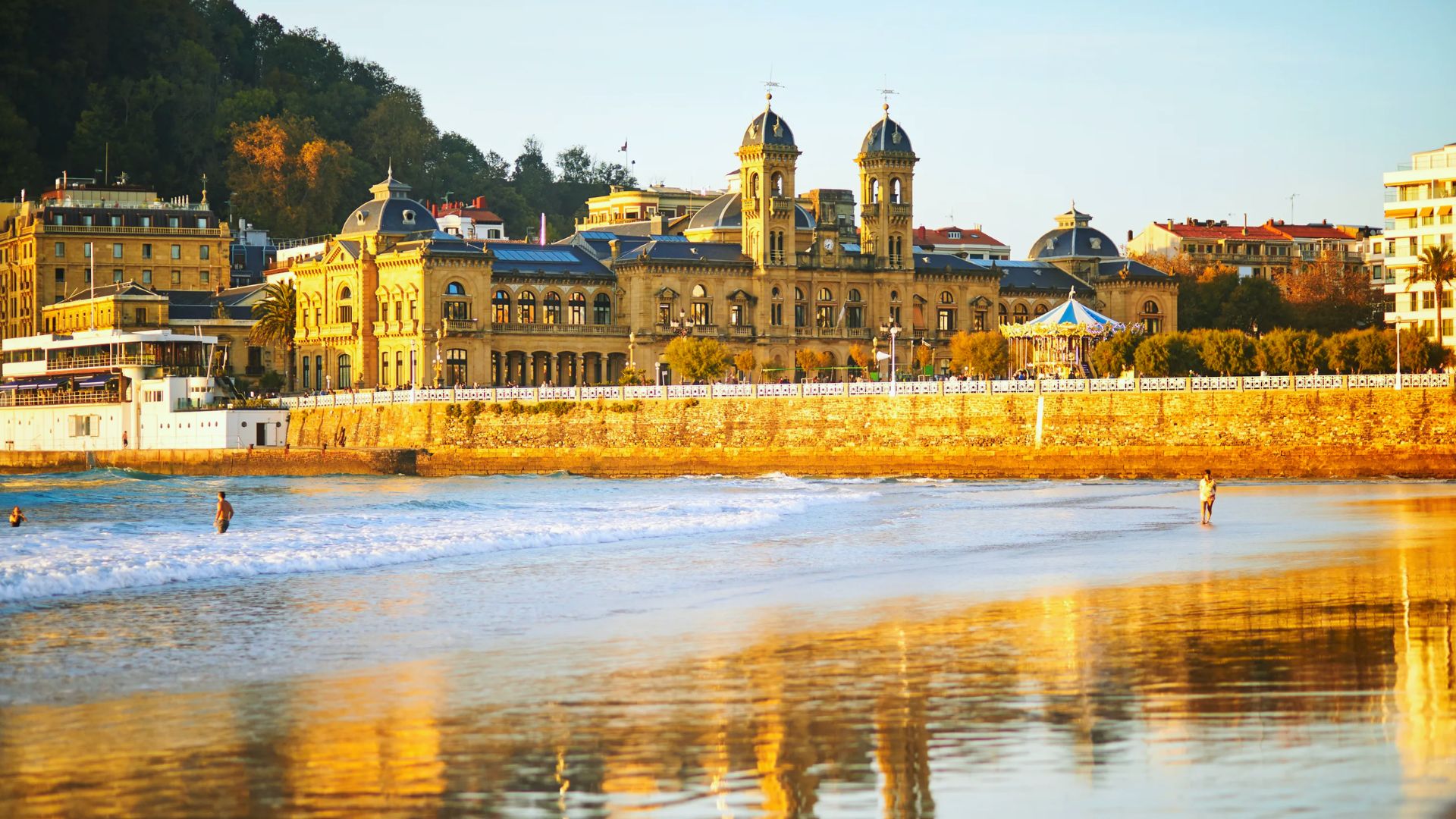 Elegant historic building with domed towers at sunset, reflecting on a sandy beach. People stroll along the shore, creating a serene, golden scene.