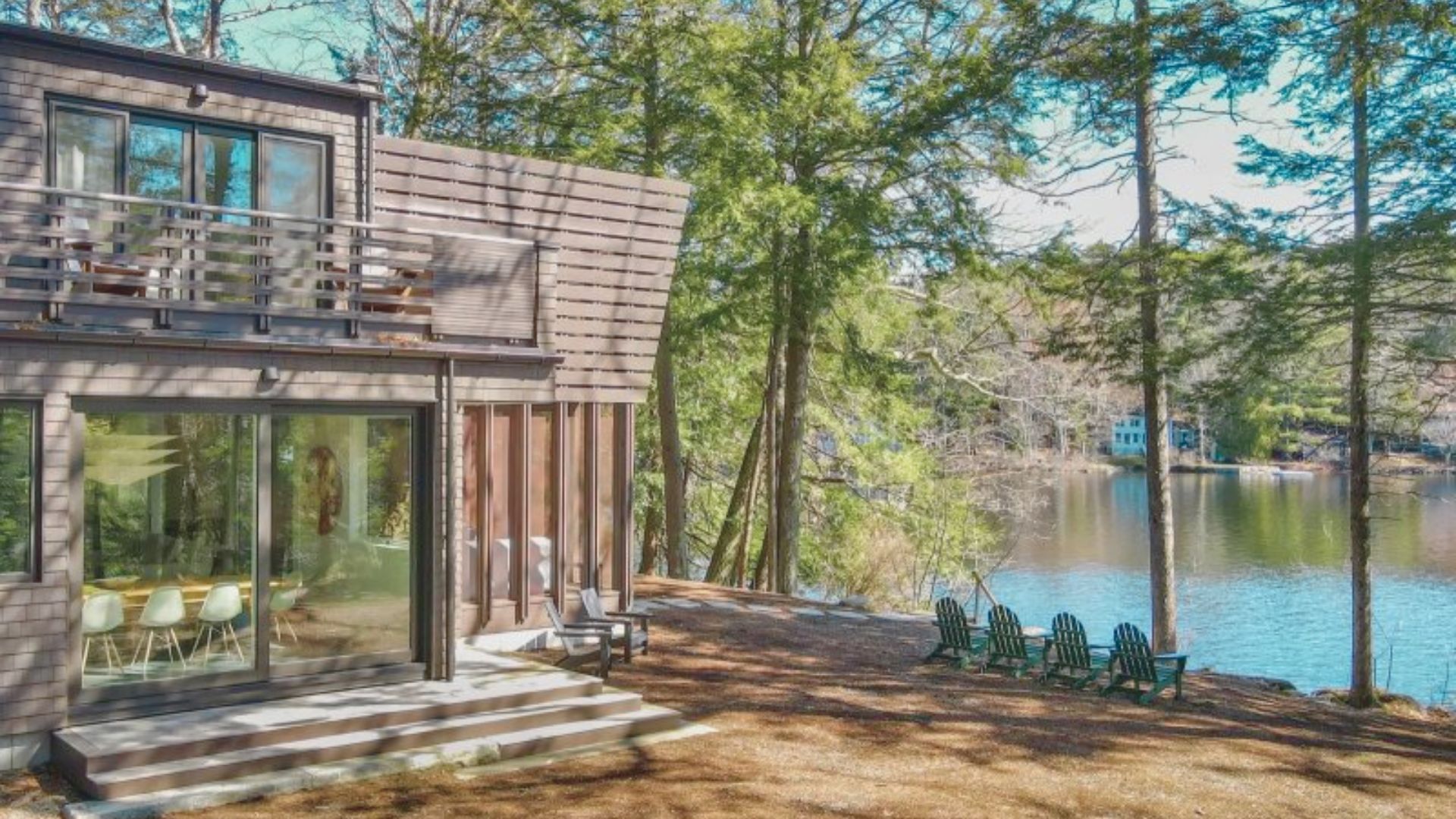 Modern lakeside cabin with large windows, surrounded by tall trees. Six green chairs are arranged facing the serene lake. Peaceful and scenic setting.