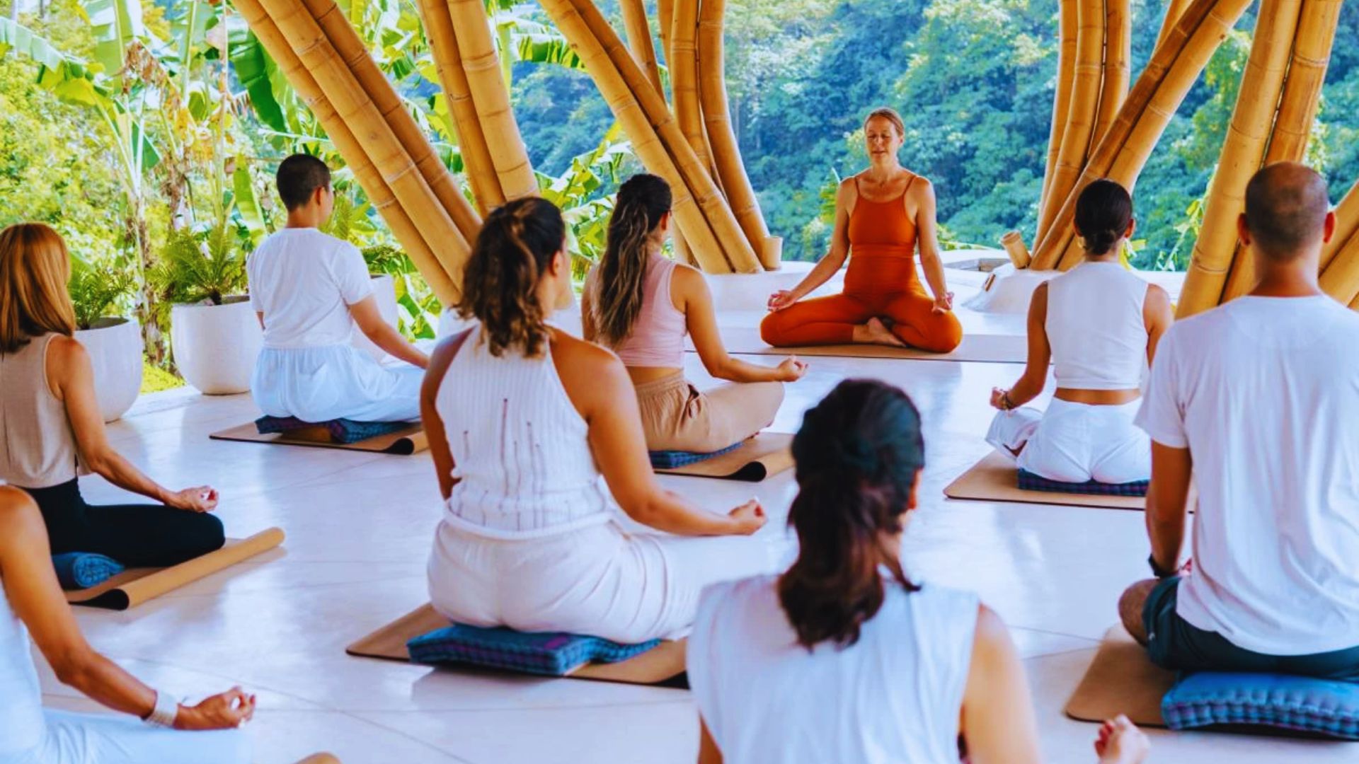 A group yoga class is seated on mats, meditating under a bamboo structure. The serene setting includes lush greenery in the background, conveying calmness.