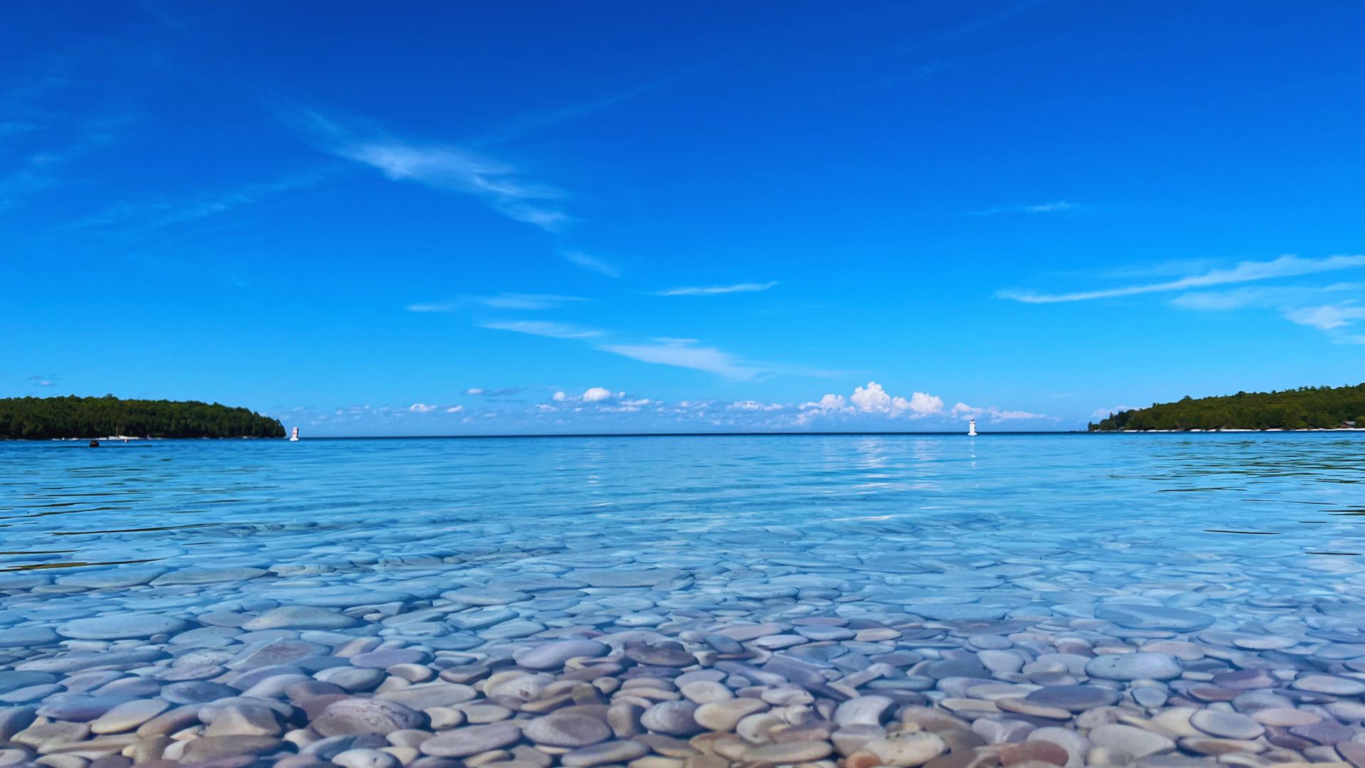 Clear blue water with smooth stones at the bottom stretches to a horizon of green islands under a vibrant blue sky, creating a serene and peaceful scene.