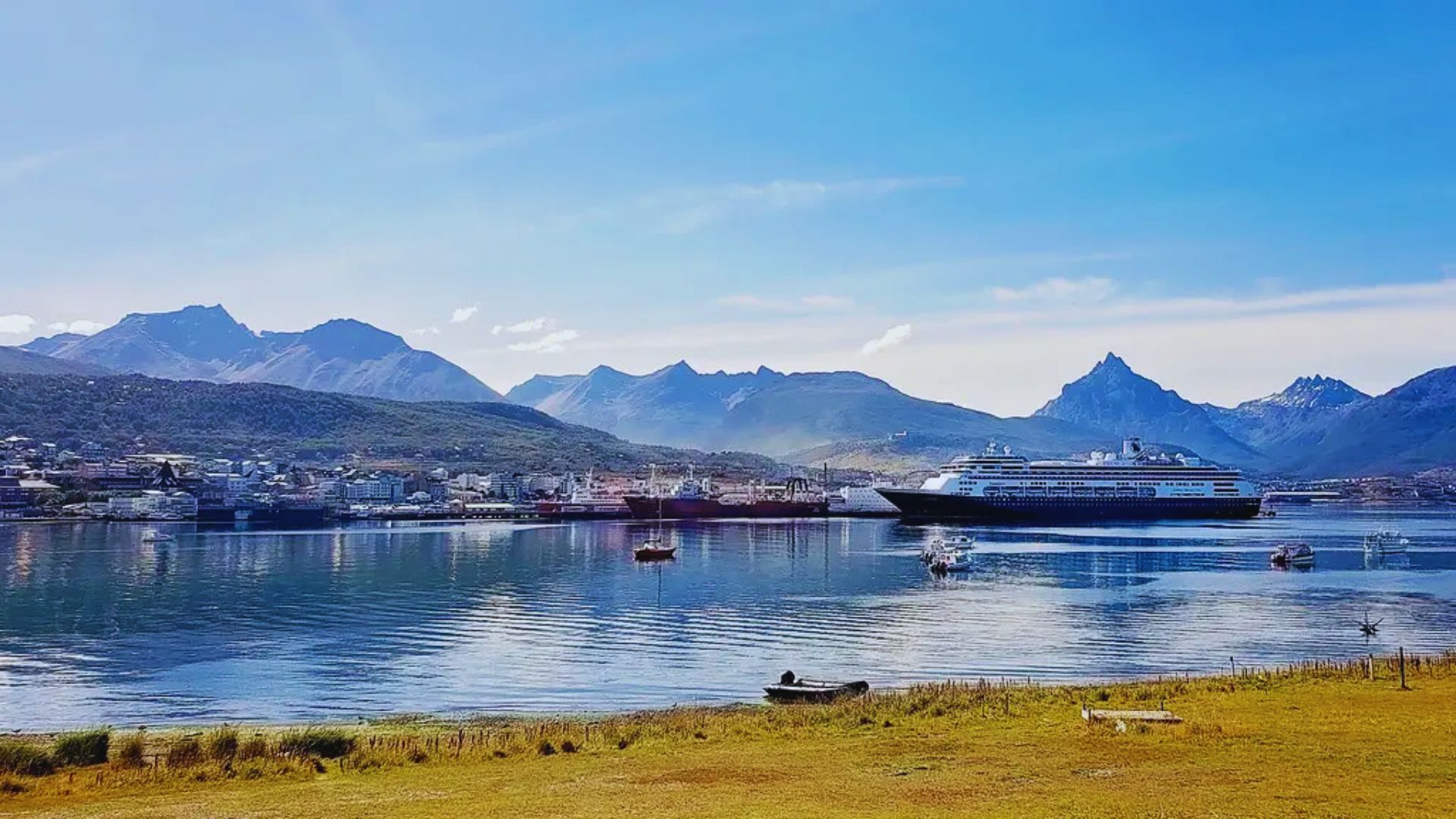 Coastal landscape with a cruise ship in a calm bay, surrounded by mountains under a clear blue sky. Peaceful scene with a small boat nearby.