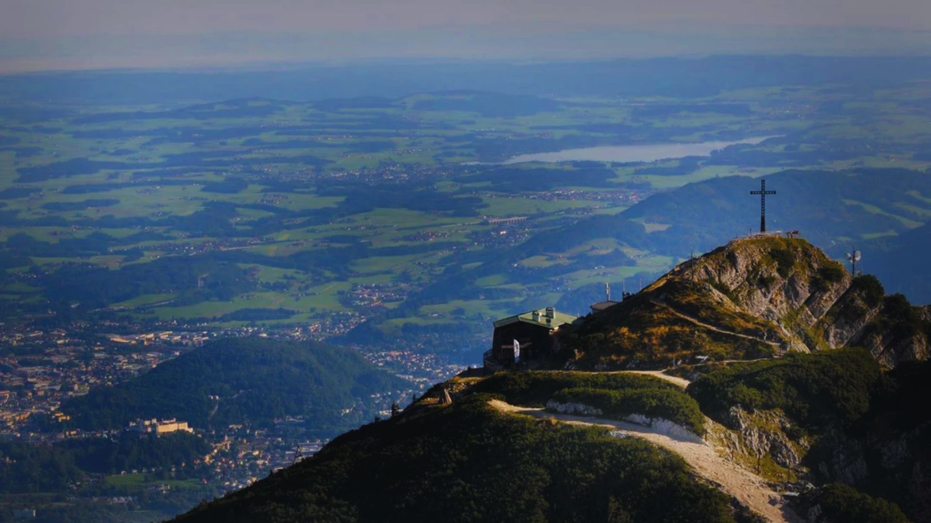 A cross atop a mountain, overlooking a vast valley below, with clear skies in the background.
