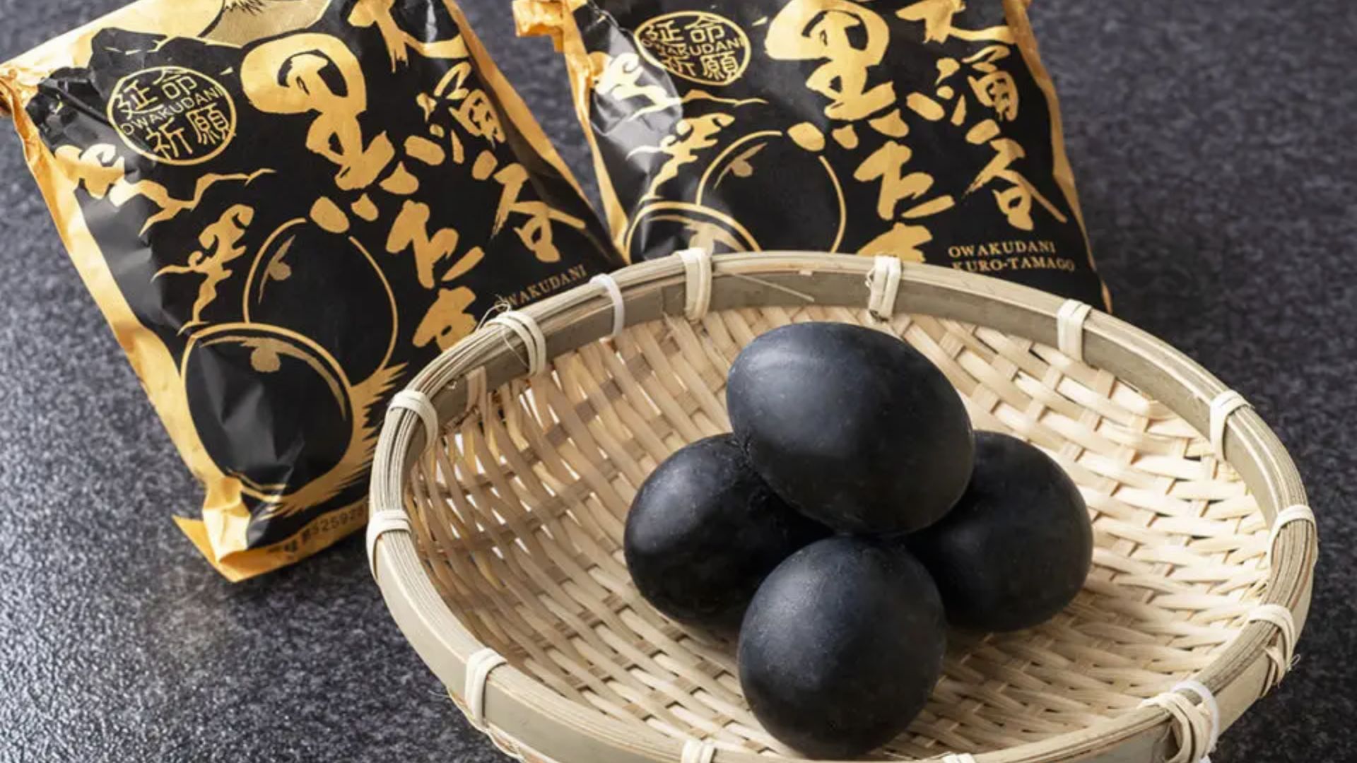 A bamboo basket holds four black eggs, with two packets in the background featuring gold and black designs, set on a textured dark surface.