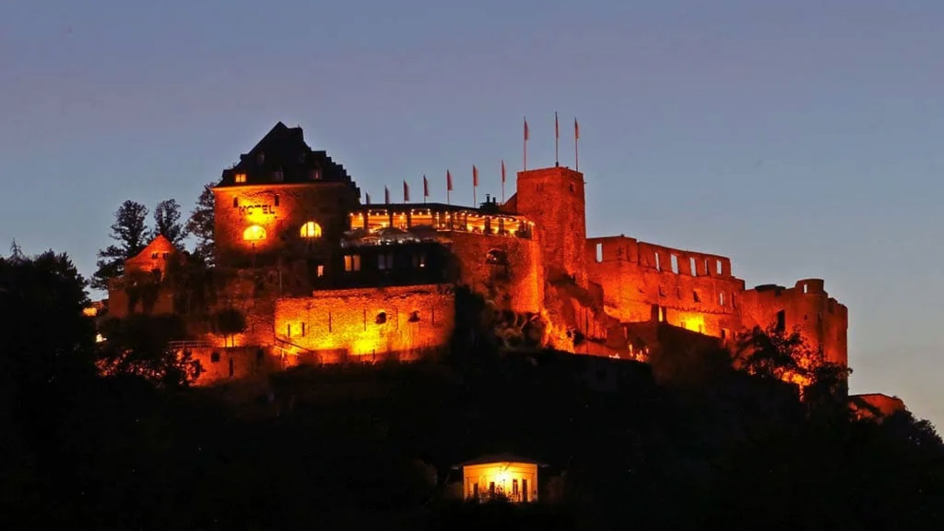 A majestic castle illuminated with warm orange lights against a twilight sky. Flags line the battlements, creating a dramatic and historic ambiance.
