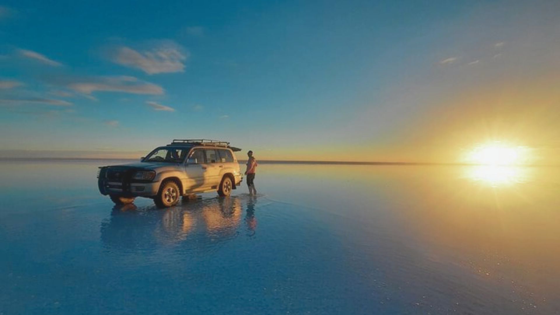 A silver SUV and a person stand on a reflective, water-covered surface under a vibrant sunset, with a calm, vast horizon and a clear, blue sky.