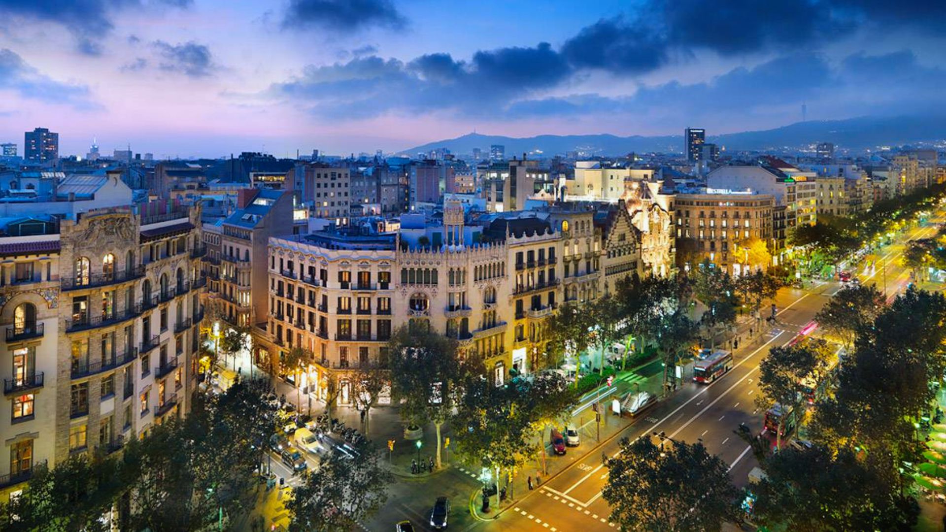 Aerial view of a vibrant cityscape at dusk in Barcelona, showcasing ornate historic buildings, tree-lined streets, and bustling evening traffic.
