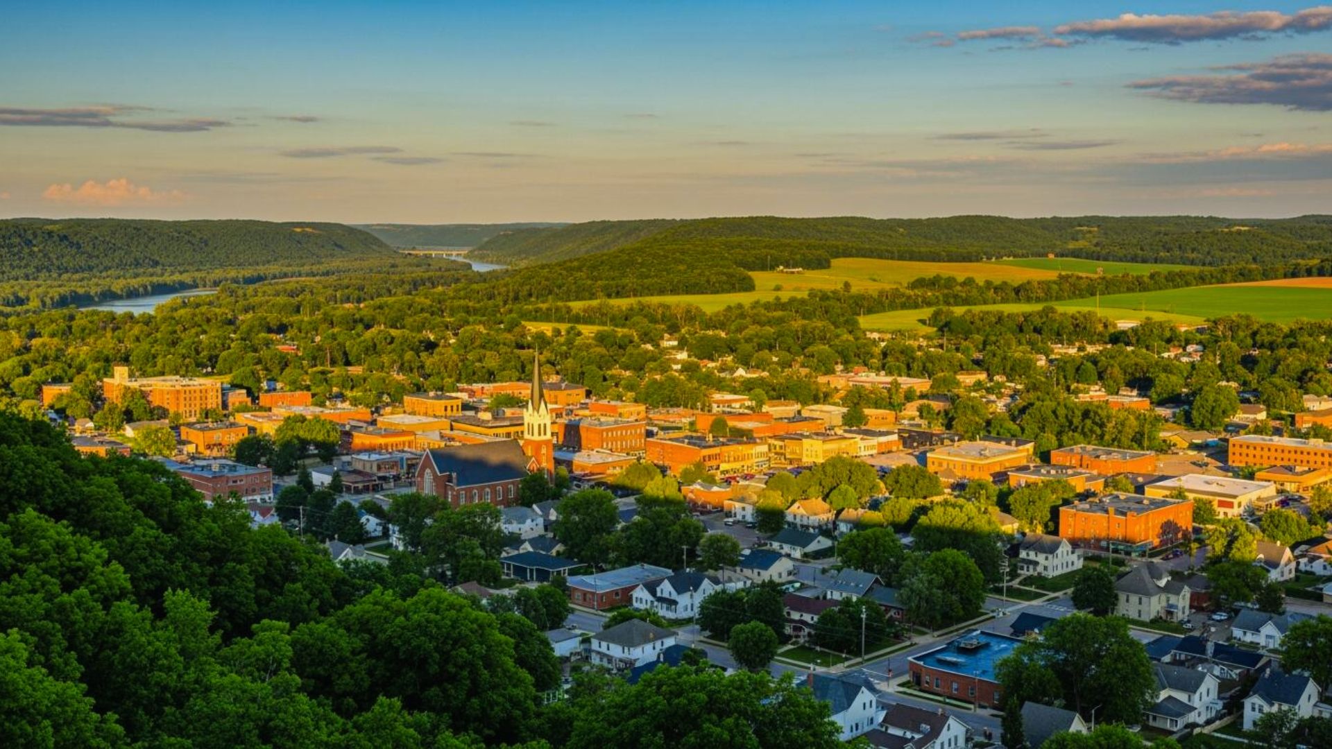 Aerial view of a small town bathed in warm sunset light, surrounded by lush green hills and fields. Central church steeple, cozy homes, and serene ambiance.