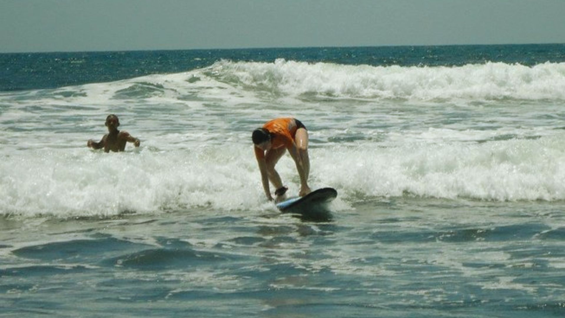 Surfer in orange top attempts to stand on a surfboard in wavy ocean water, while a swimmer watches nearby under a clear blue sky.