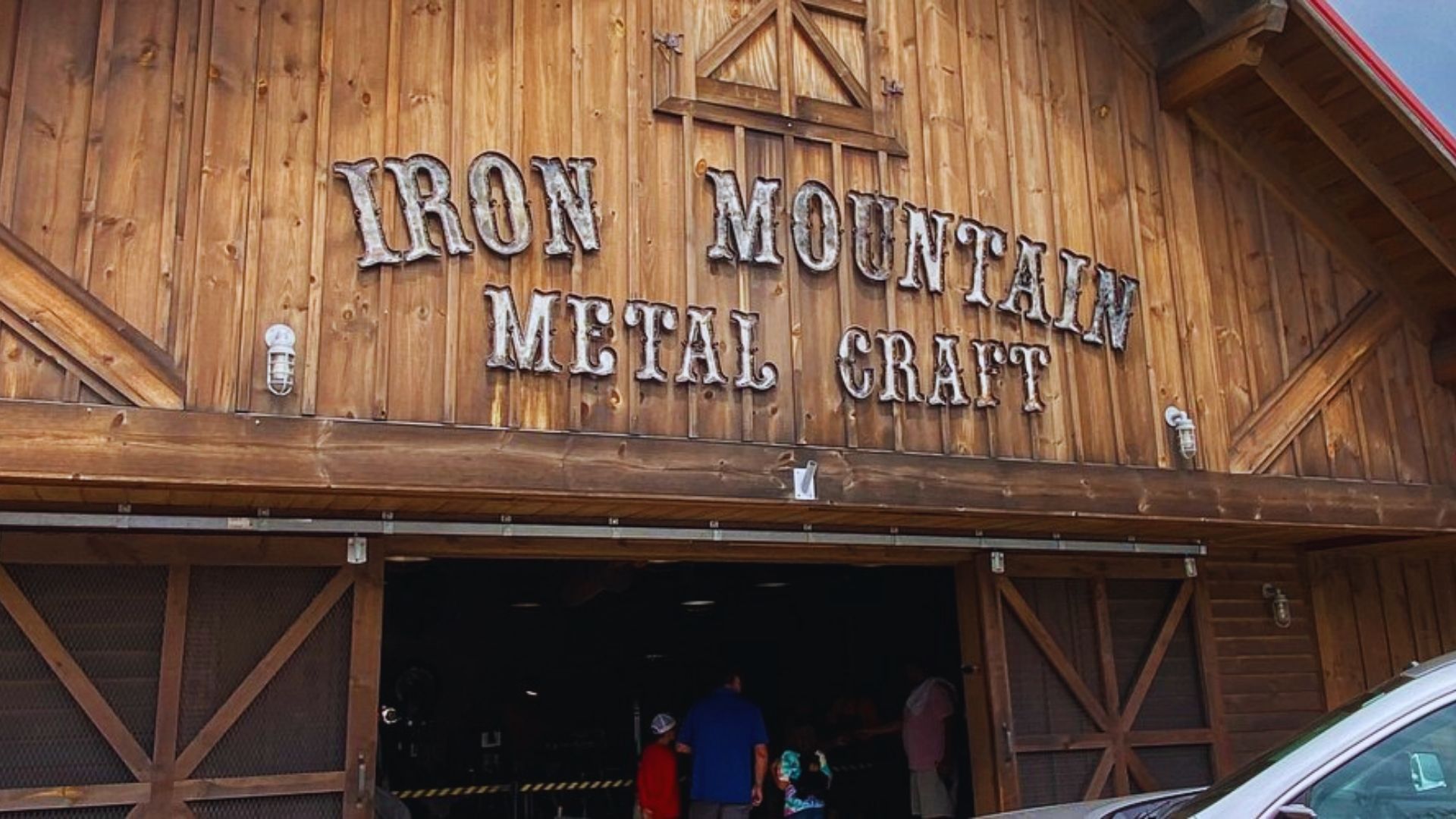 Wooden barn-style building with "Iron Mountain Metal Craft" sign. Entrance features people entering, exuding a rustic, welcoming atmosphere.