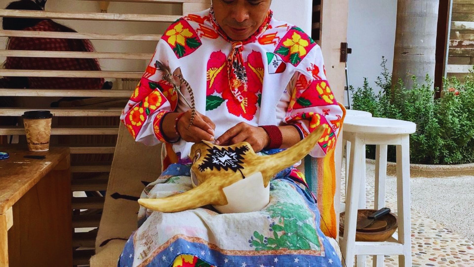 A person in colorful, traditional attire intricately paints a carved object, focusing intently. The setting is cozy and creative, conveying dedication.