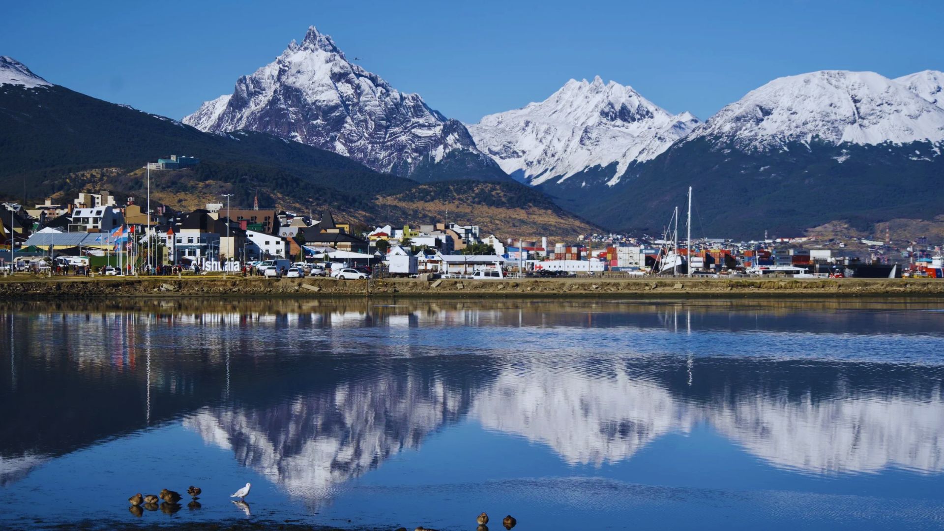 A picturesque town sits by a calm lake, reflecting snow-capped mountains under a clear blue sky, exuding a serene and tranquil atmosphere.