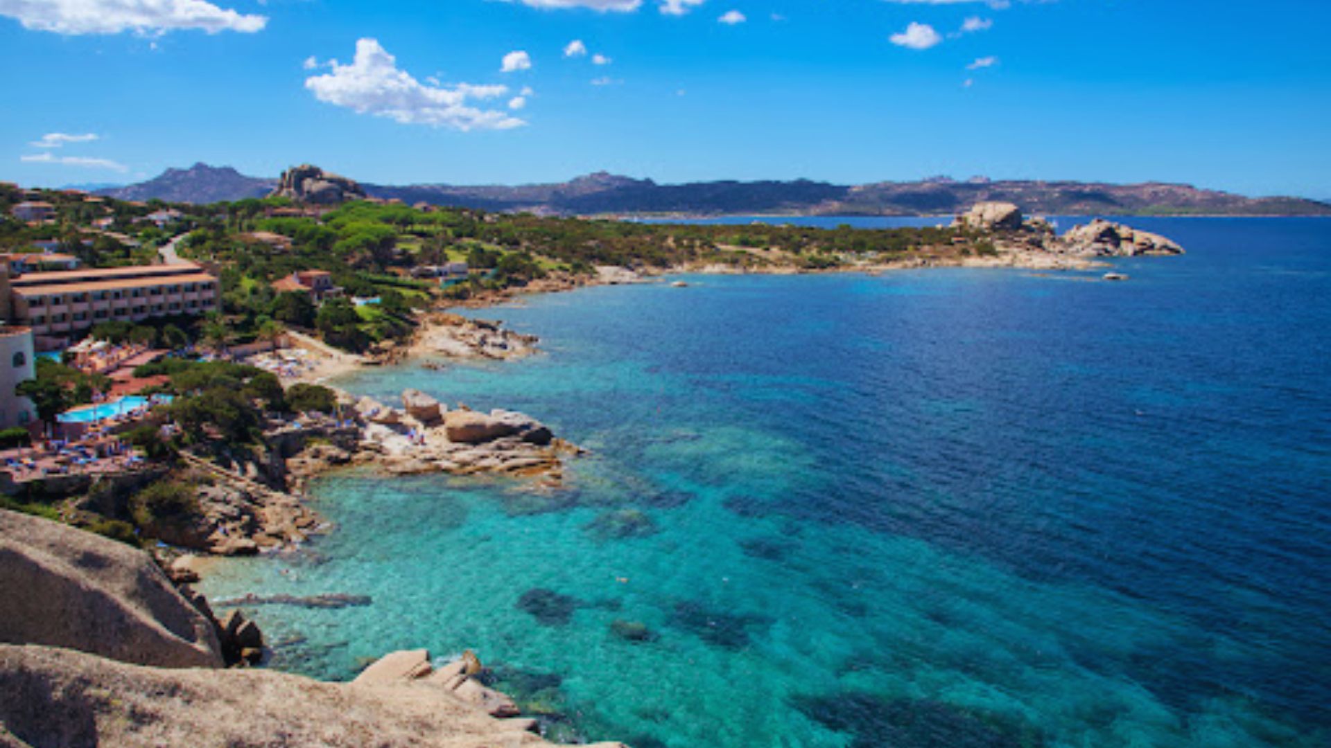 Coastal landscape featuring turquoise waters, rocky shoreline, and lush green vegetation. Buildings overlook the scenic bay, under a bright blue sky.