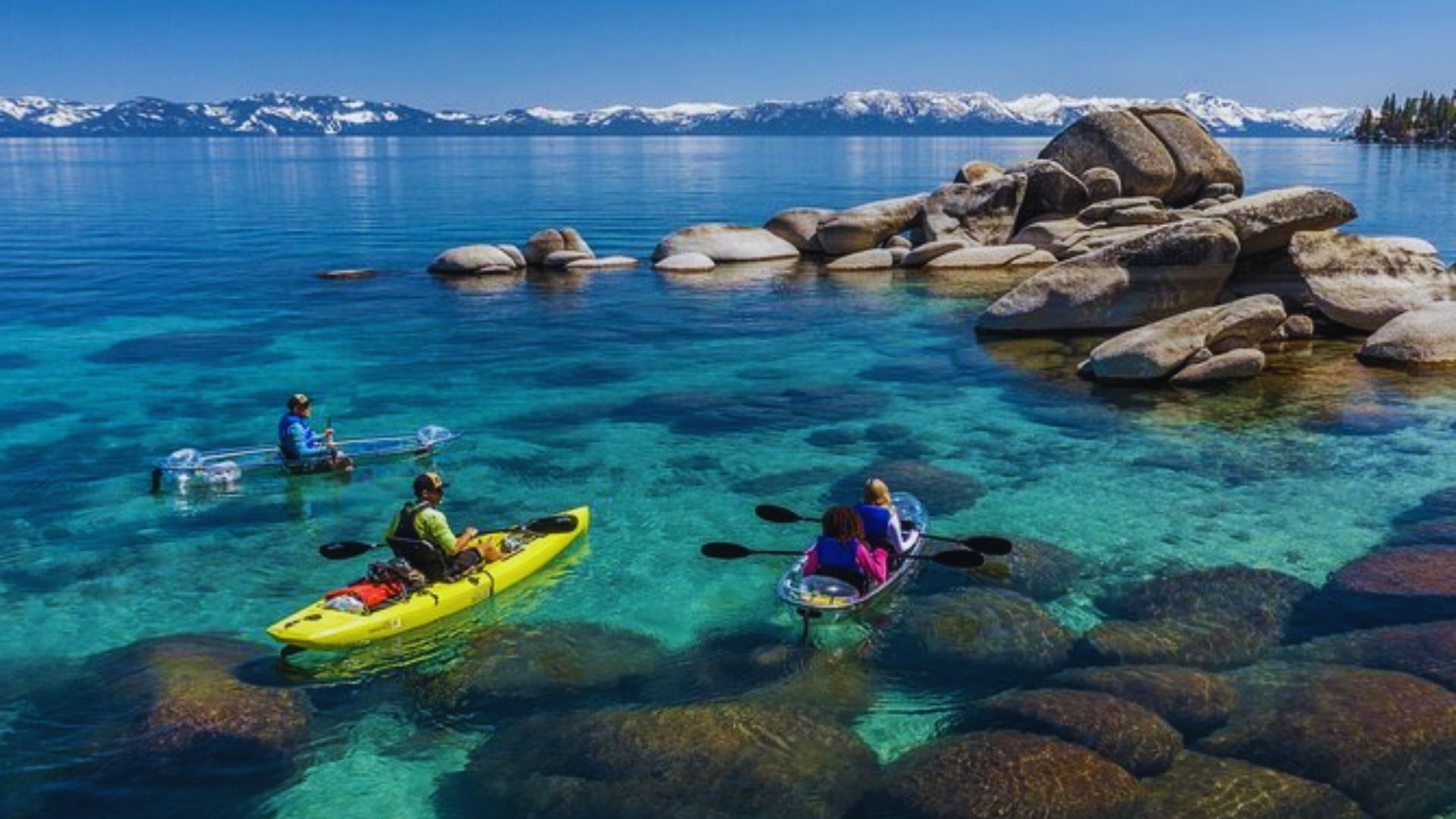 Three people kayaking in clear water, surrounded by majestic mountains under a bright blue sky.