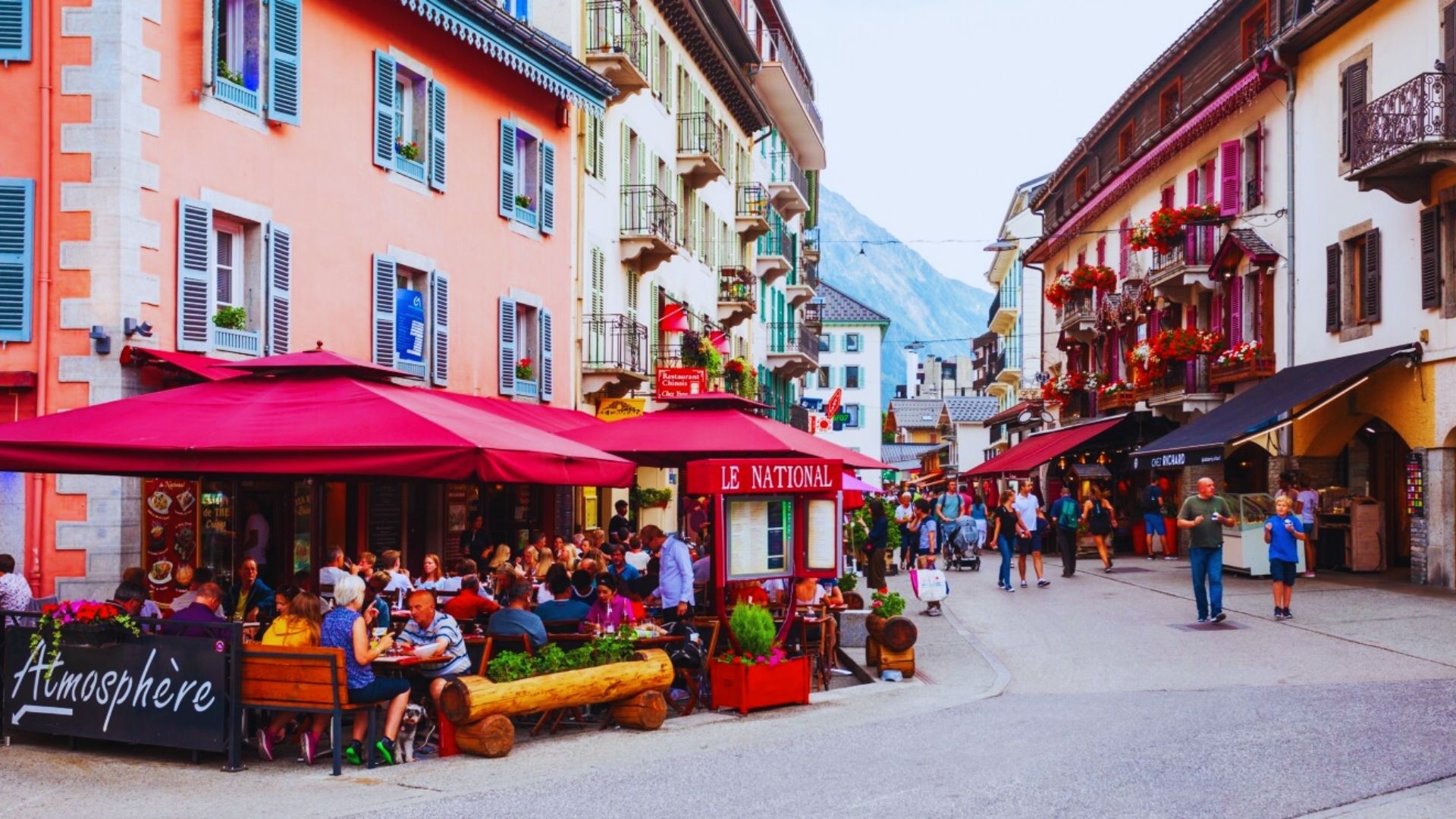 A bustling street scene with numerous people dining outside various restaurants under sunny weather.