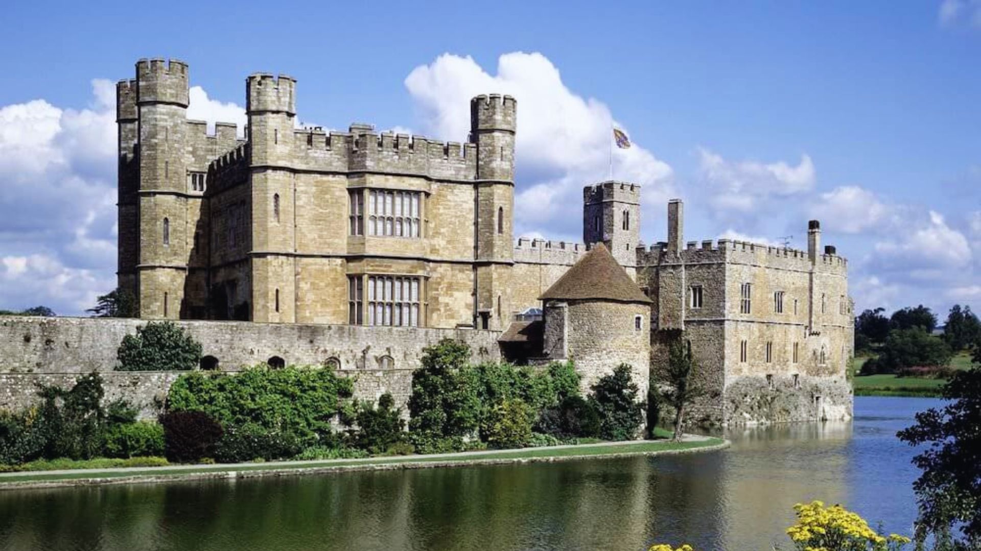 Majestic stone castle with towers, surrounded by a moat and lush greenery. A partly cloudy sky adds a serene backdrop. Historic and tranquil ambiance.