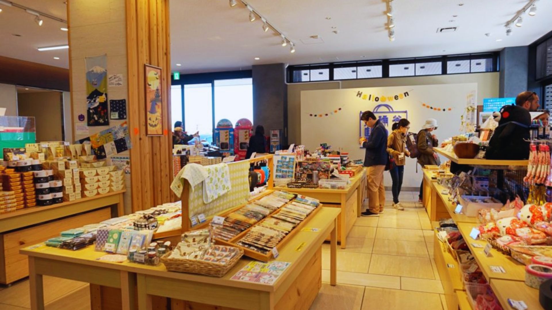 A brightly lit shop features wooden display tables with assorted snacks and souvenirs. People browse near a wall with "Halloween" decor, creating a festive atmosphere.