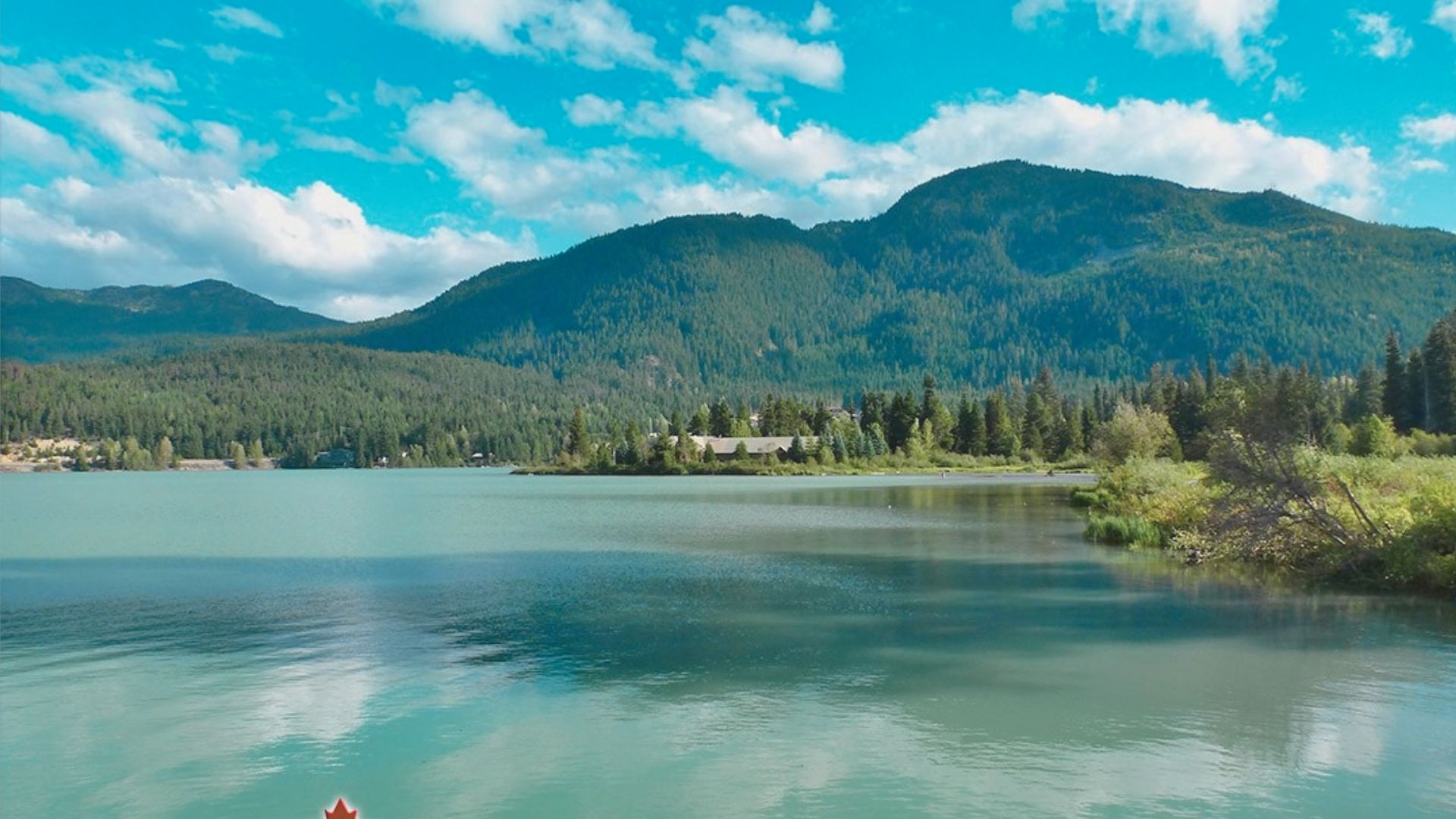 A serene lake reflects blue skies and fluffy clouds, surrounded by lush green forests and distant mountains, evoking a peaceful, natural tranquility.