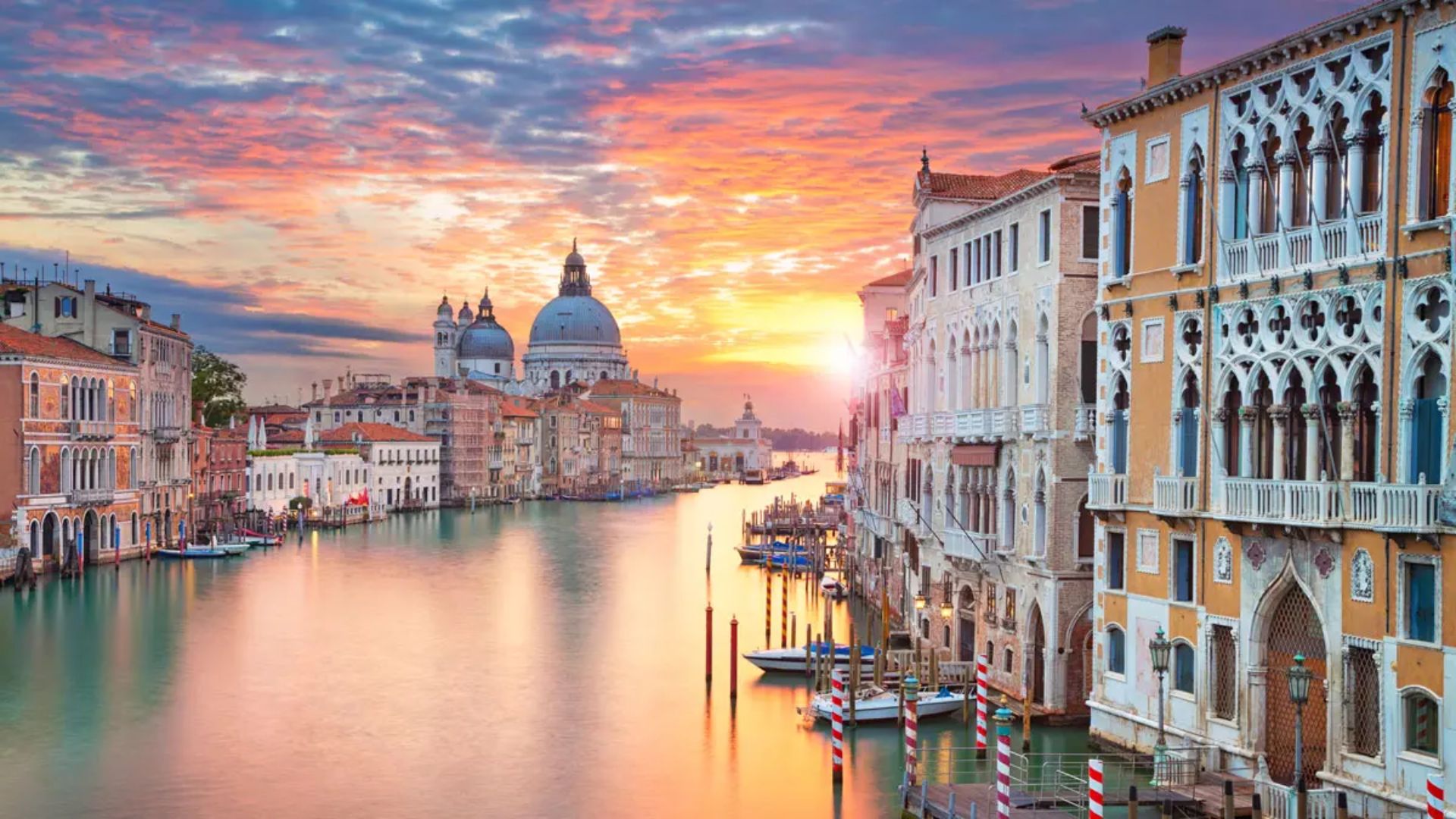 Venice at sunset with a vibrant sky above the Grand Canal. Historic buildings line the water; boats float gently. A serene, picturesque scene.