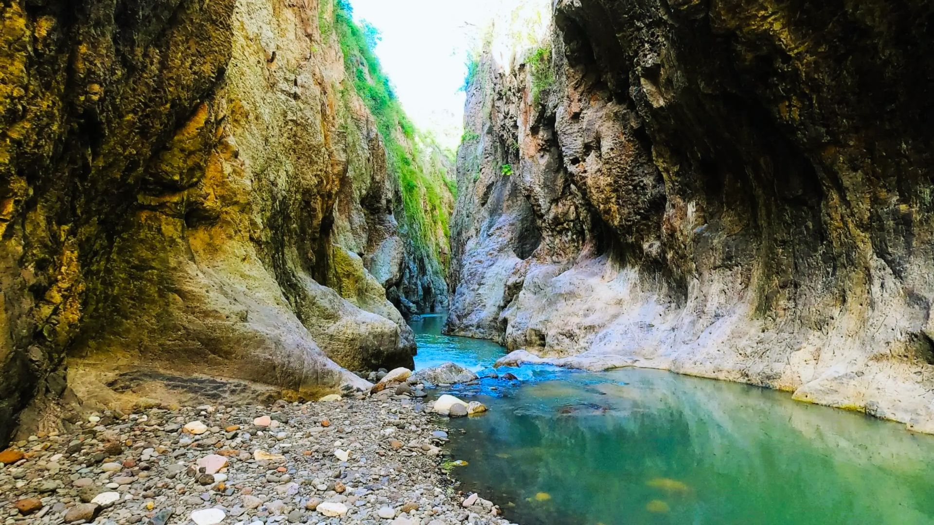 A serene narrow canyon with rugged, towering rock walls. A turquoise stream flows through the rocky landscape under a bright sky, creating a peaceful vibe.