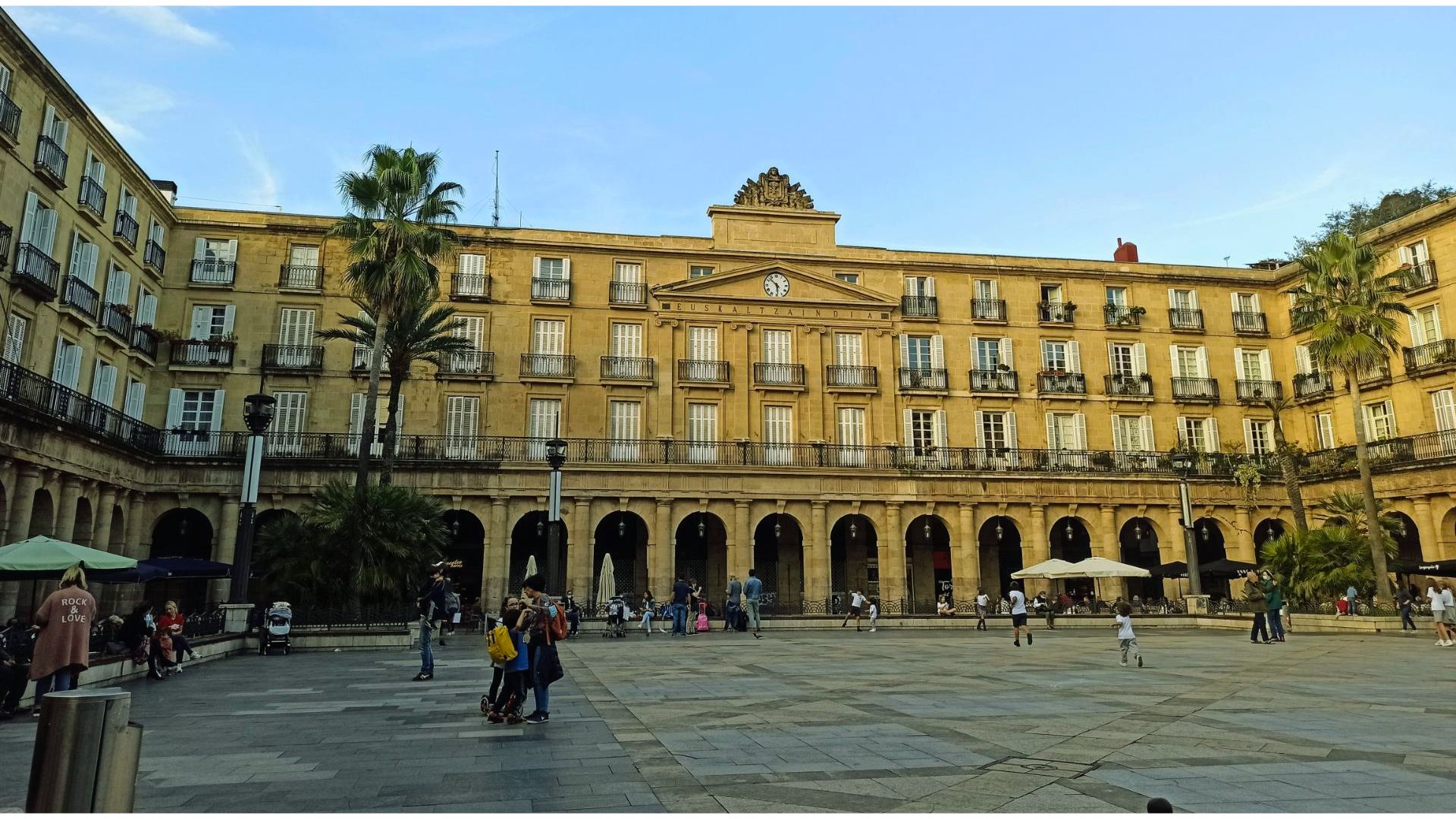A large, historic building with arches and balconies surrounds a spacious plaza. People stroll, and palm trees create a lively, relaxed atmosphere.