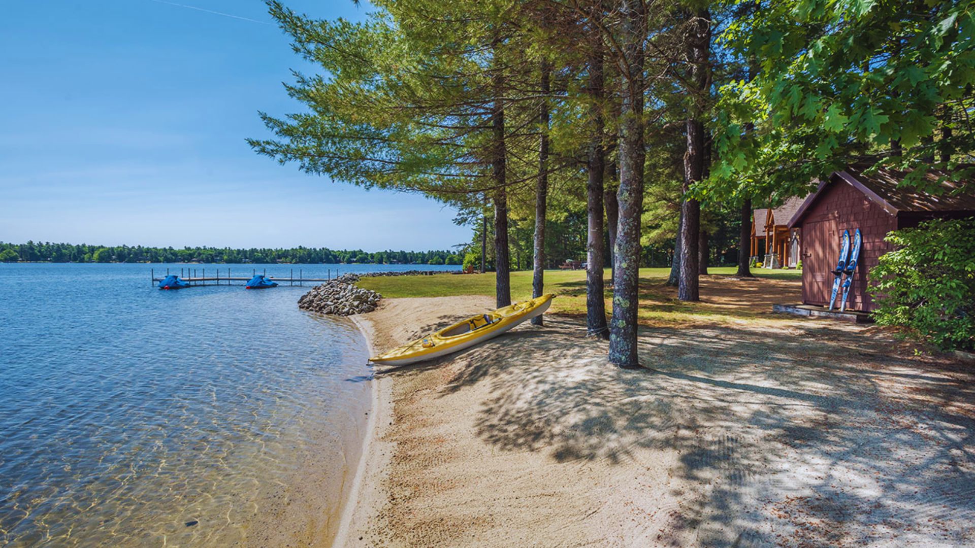 A serene lakeside scene with a sandy beach, yellow kayak, and paddleboats on calm water. Trees provide shade near a wooden cabin, evoking tranquility.