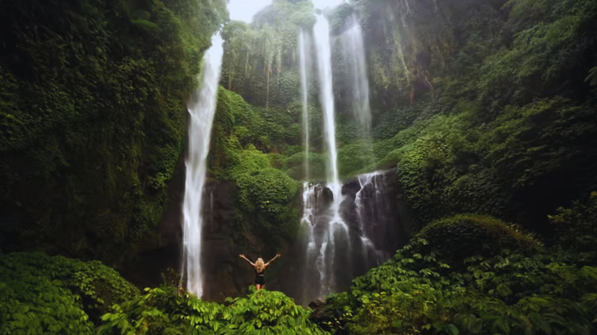 A person stands with arms raised in front of a majestic, lush green waterfall. Sunlight filters through the foliage, creating a serene, awe-inspiring scene.