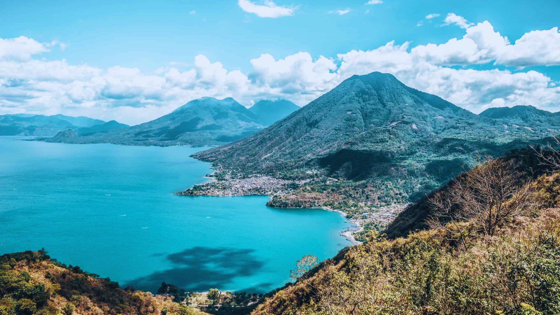 Scenic view of a turquoise lake with lush, green mountains under a vibrant blue sky with fluffy clouds, conveying serenity and natural beauty.
