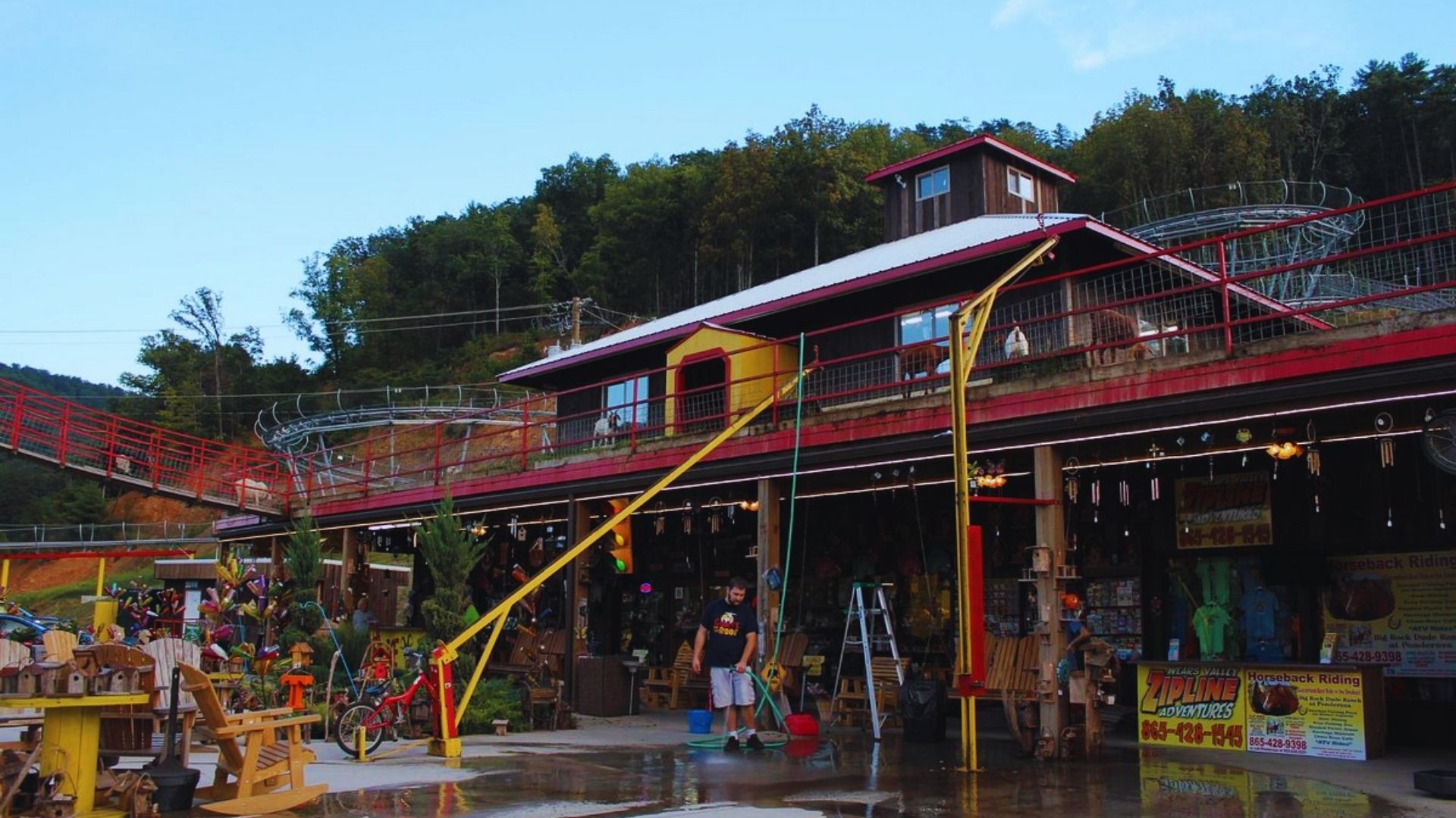Outdoor wooden shop with crafts and furniture under a red-roofed building. A mountain coaster track winds above. Lush green hills in the background.