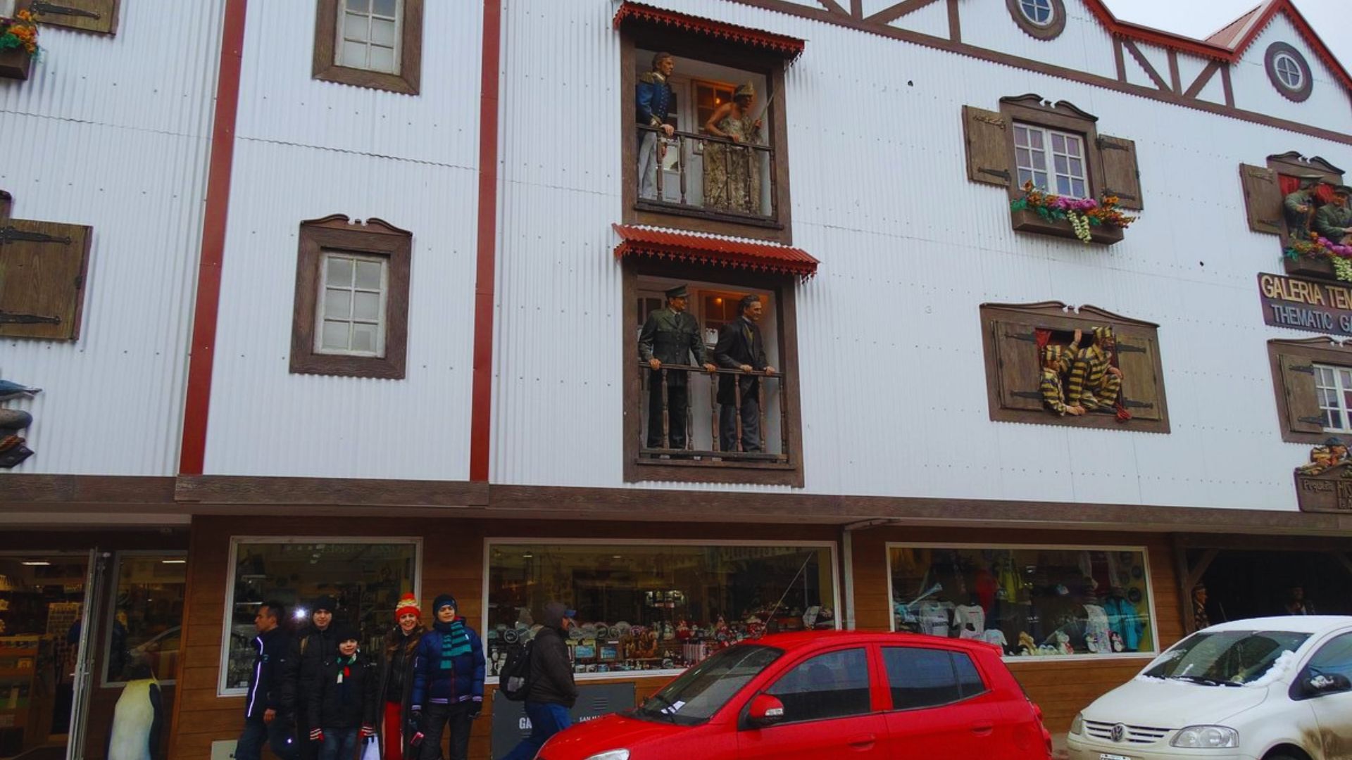 Street scene with a Bavarian-style building featuring wooden shutters and mannequin figures on a balcony. People walk past below, and cars are parked nearby.