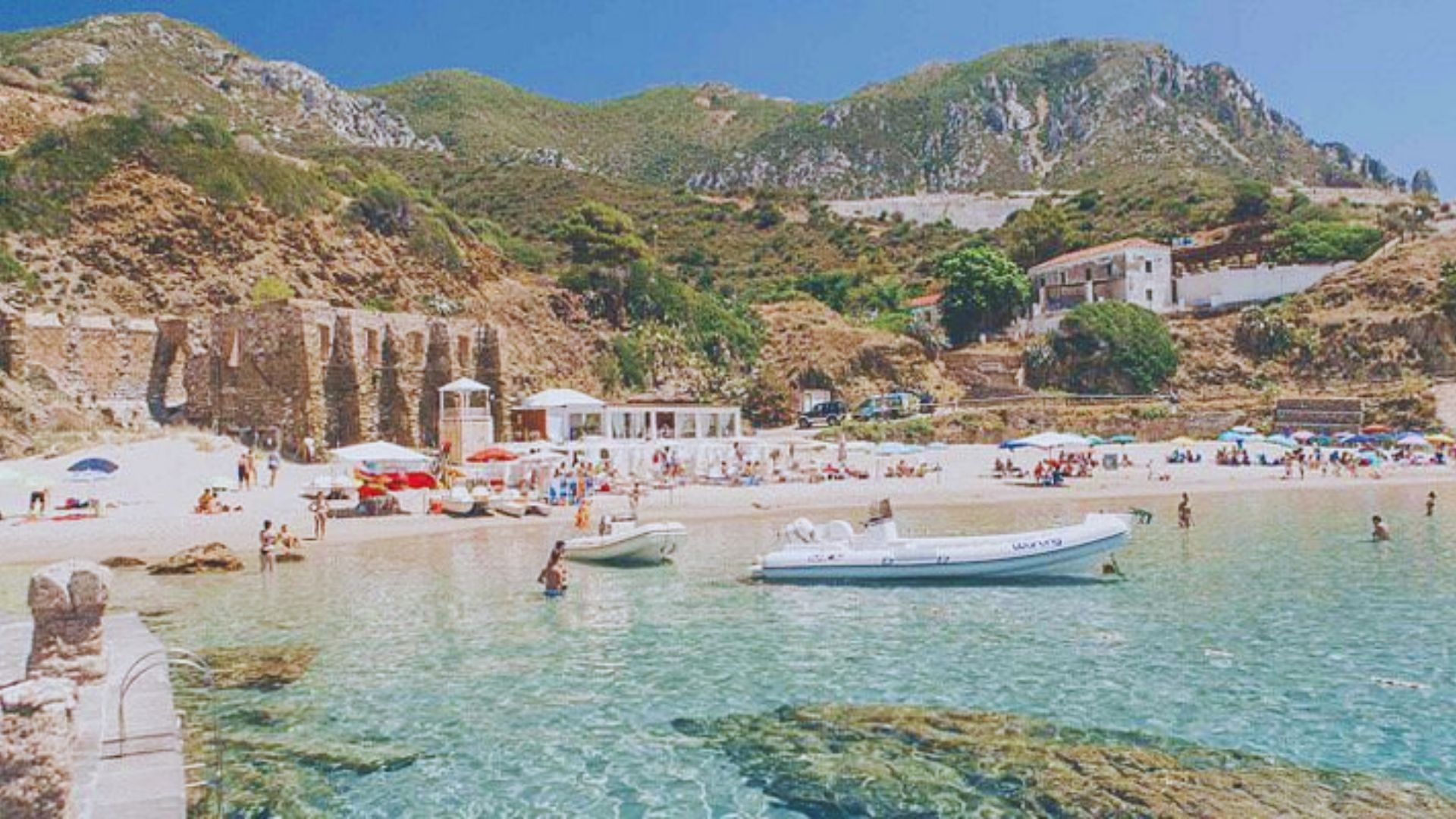 Sunny beach scene with clear turquoise water, rocky shoreline, and lush green hills. People enjoy the beach, boats float nearby, and umbrellas dot the sand.