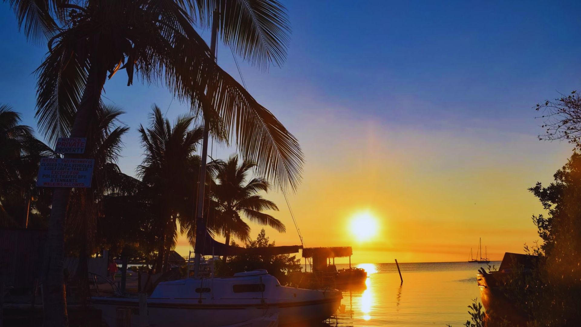 A vibrant sunset over water, featuring silhouetted boats and palm trees against a colorful sky.