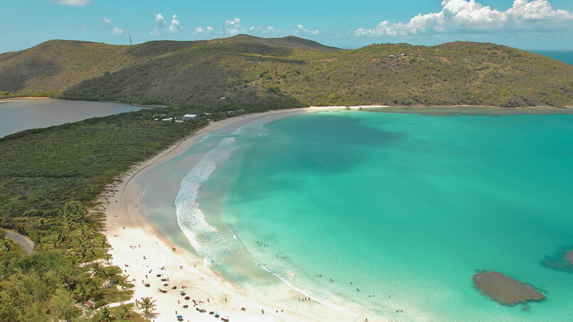 Aerial view of a tranquil beach with turquoise waters, a crescent-shaped shoreline, and surrounding hills. Sparse clouds dot the blue sky, creating a serene scene.