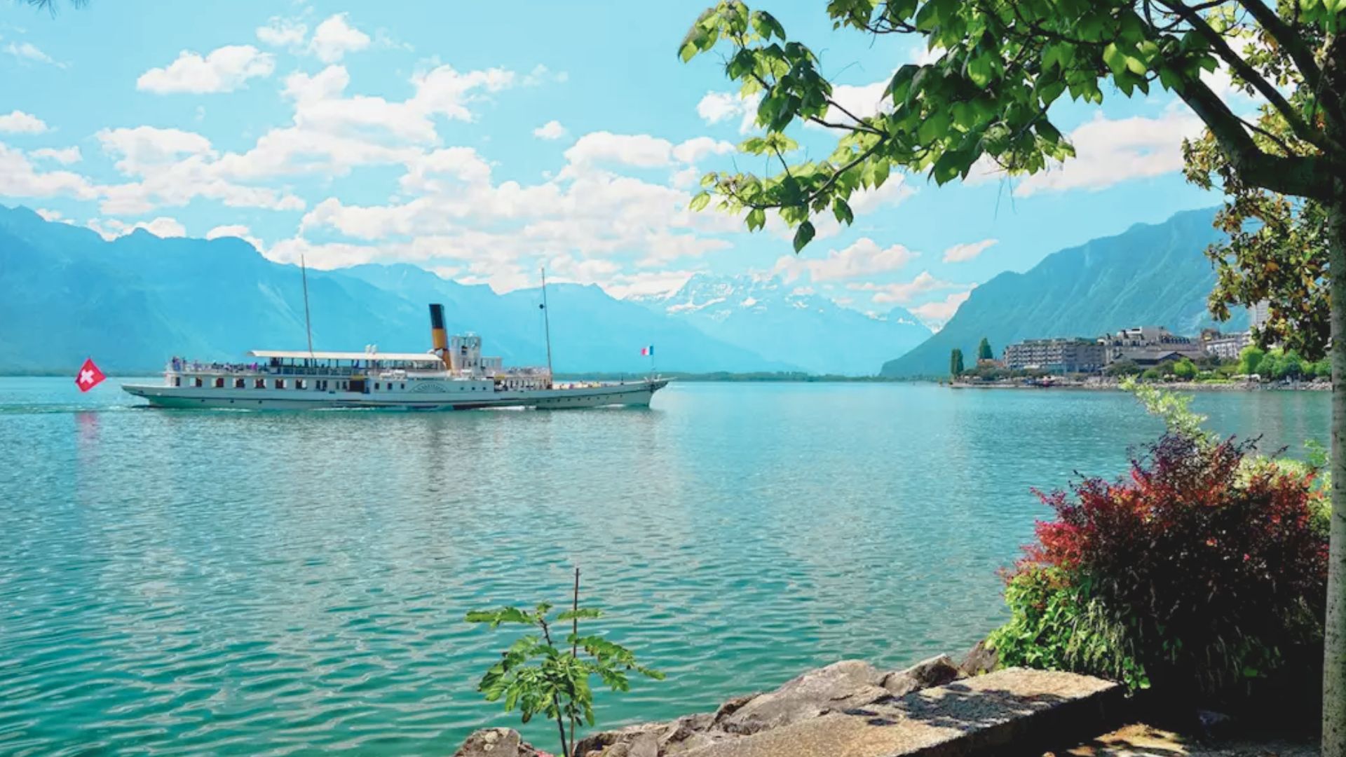 A serene lake scene with a vintage boat sailing, Swiss flag waving. Snow-capped mountains under a bright, cloudy sky. Foreground with leafy trees and colorful bushes.